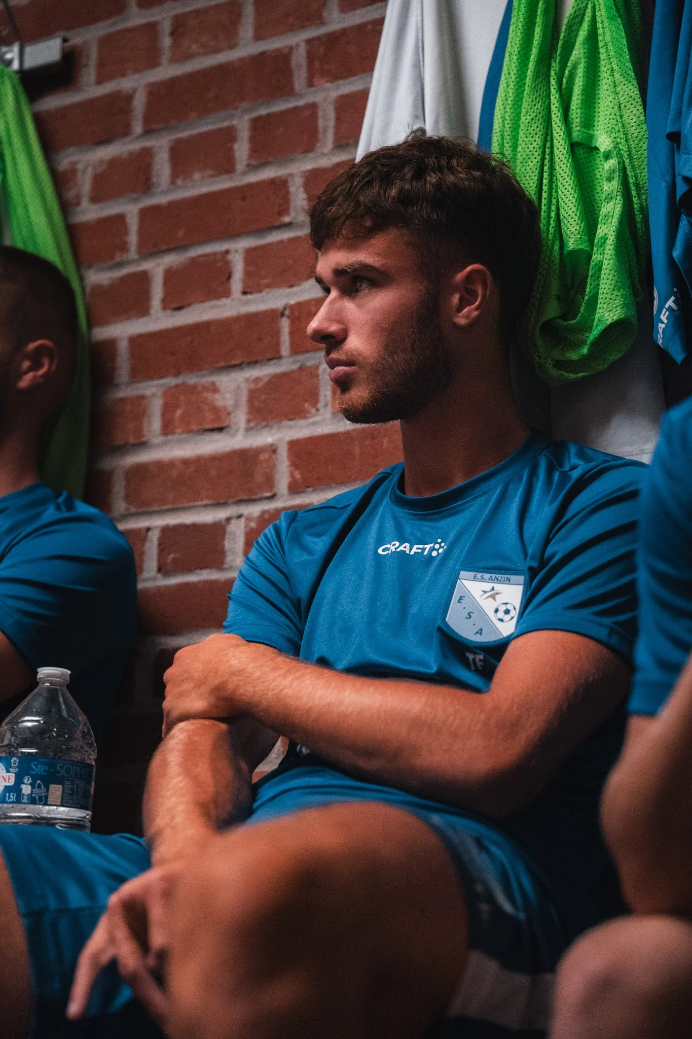 Un jeune homme dans un maillot de football bleu, assis dans une pièce avec un mur en briques rouges derrière lui, semble concentré ou pensif, avec des vêtements et une bouteille d'eau à ses côtés.