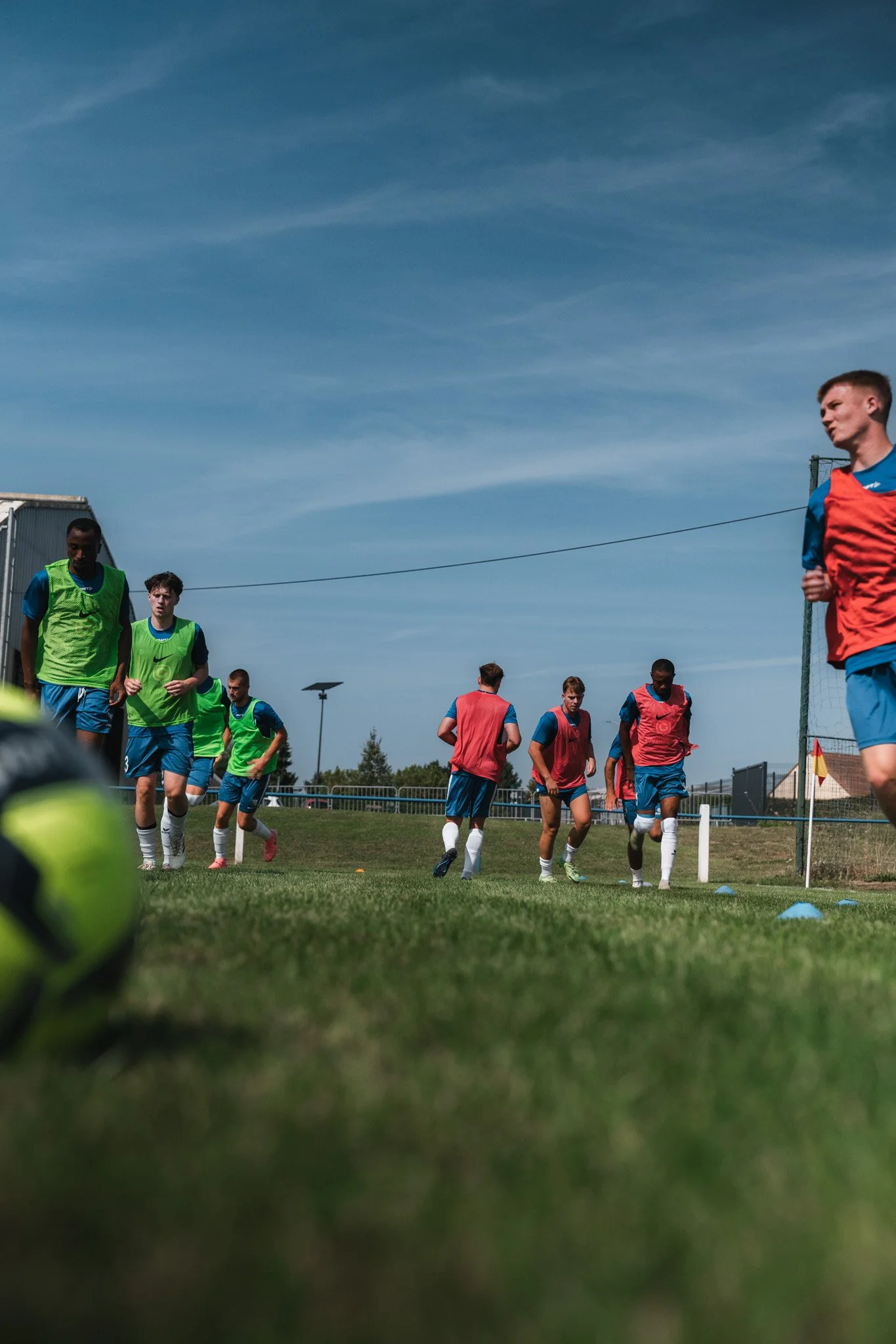 Des joueurs de football en entraînement sur un terrain en plein air, portant des maillots colorés, avec un ballon à l'avant-plan et un ciel bleu en arrière-plan.