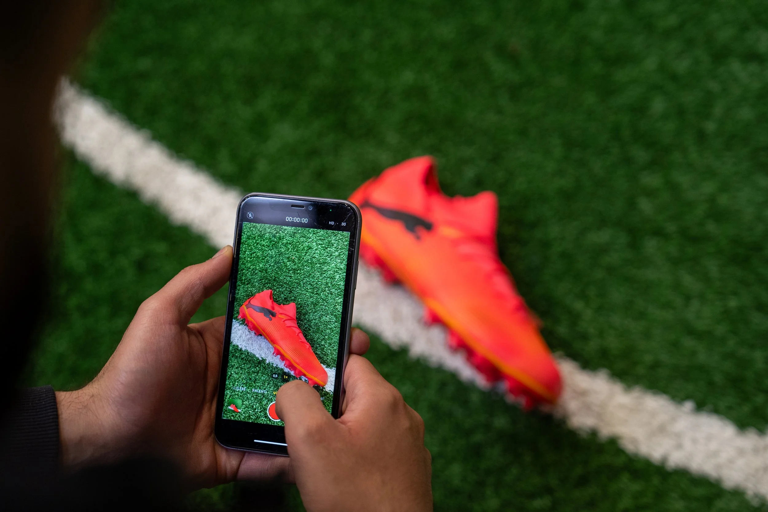 Une personne prend en photo une chaussure de sport orange sur un terrain de football en herbe, avec une ligne blanche en arrière-plan.
