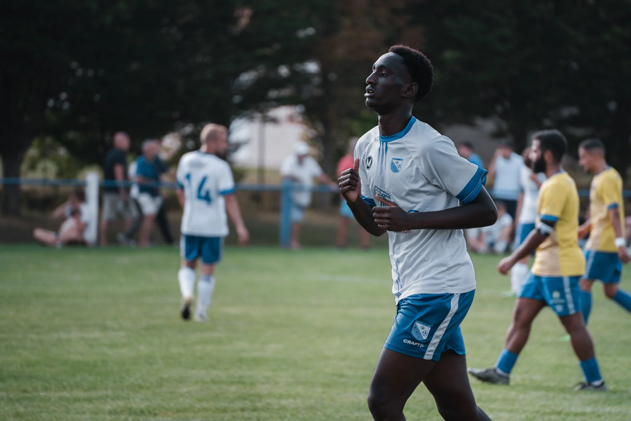 Un joueur de football en uniforme blanc et bleu, courant sur le terrain lors d'un match, avec d'autres joueurs et spectateurs en arrière-plan.