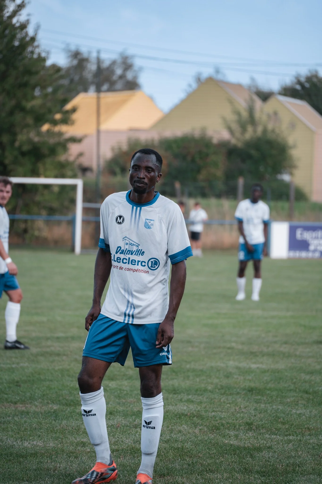 Un joueur de football en uniforme blanc et bleu sur un terrain de football, avec d'autres joueurs en arrière-plan.