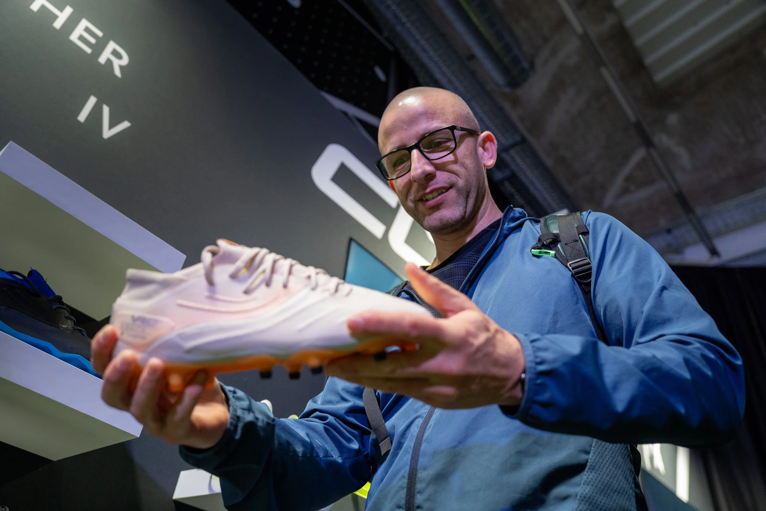 Un homme portant des lunettes et une veste examine une chaussure de course colorée en exposition.