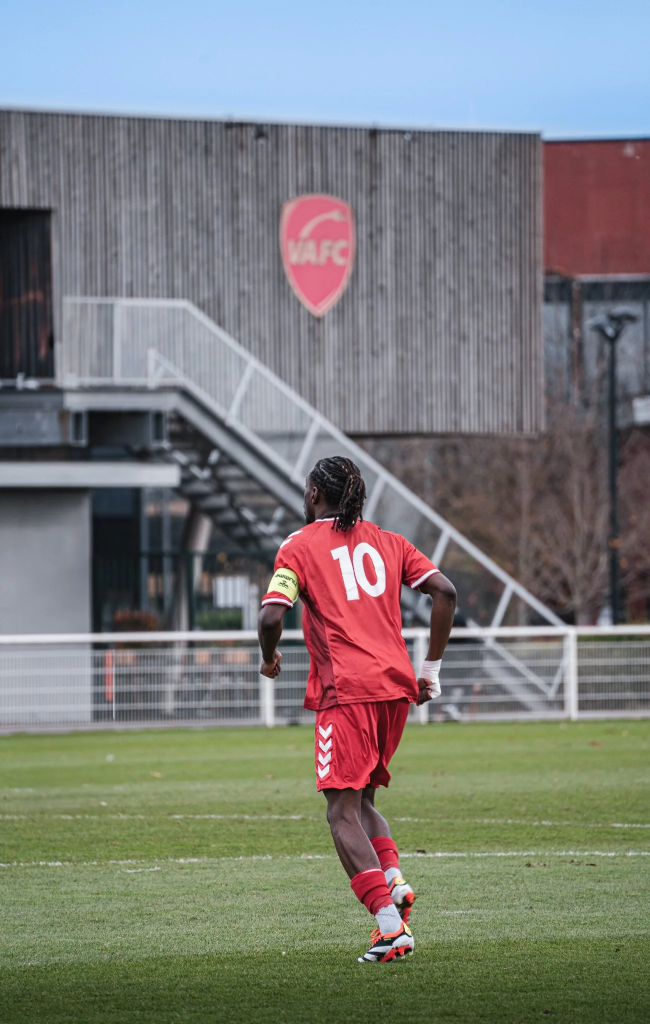 Un joueur de football vêtu d'un maillot rouge portant le numéro 10, courant sur un terrain d'herbe, avec un bâtiment en bois en arrière-plan portant un logo de club de football