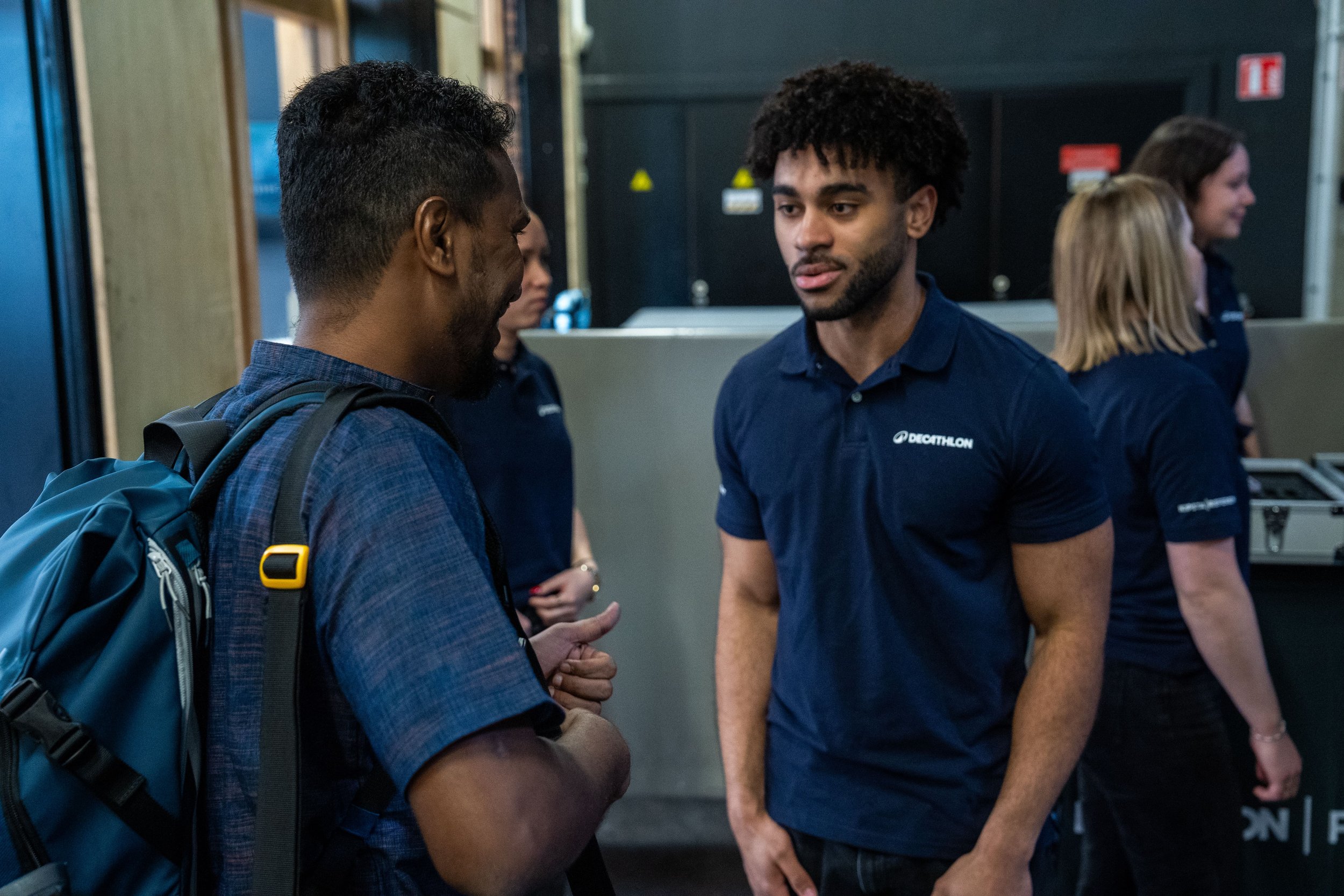Deux hommes discutent dans un environnement intérieur, l'un avec un sac à dos. Ils portent tous deux des polo bleu avec le logo Décathlon.
