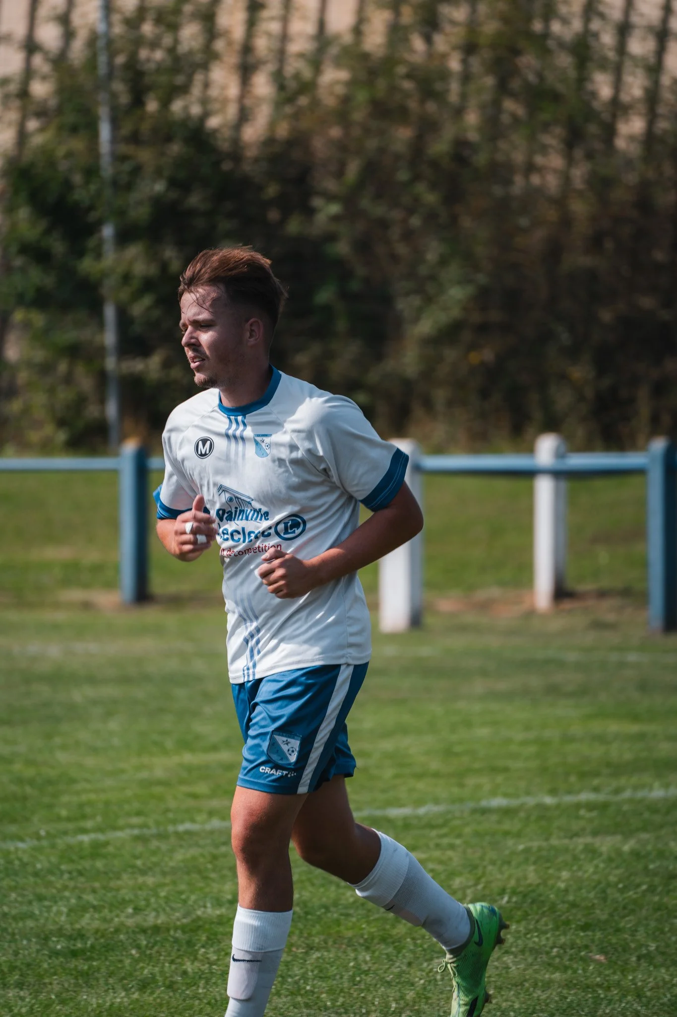 Un joueur de football en train de courir sur le terrain, portant un maillot blanc et bleu, avec un fond de clôture et d'arbres.