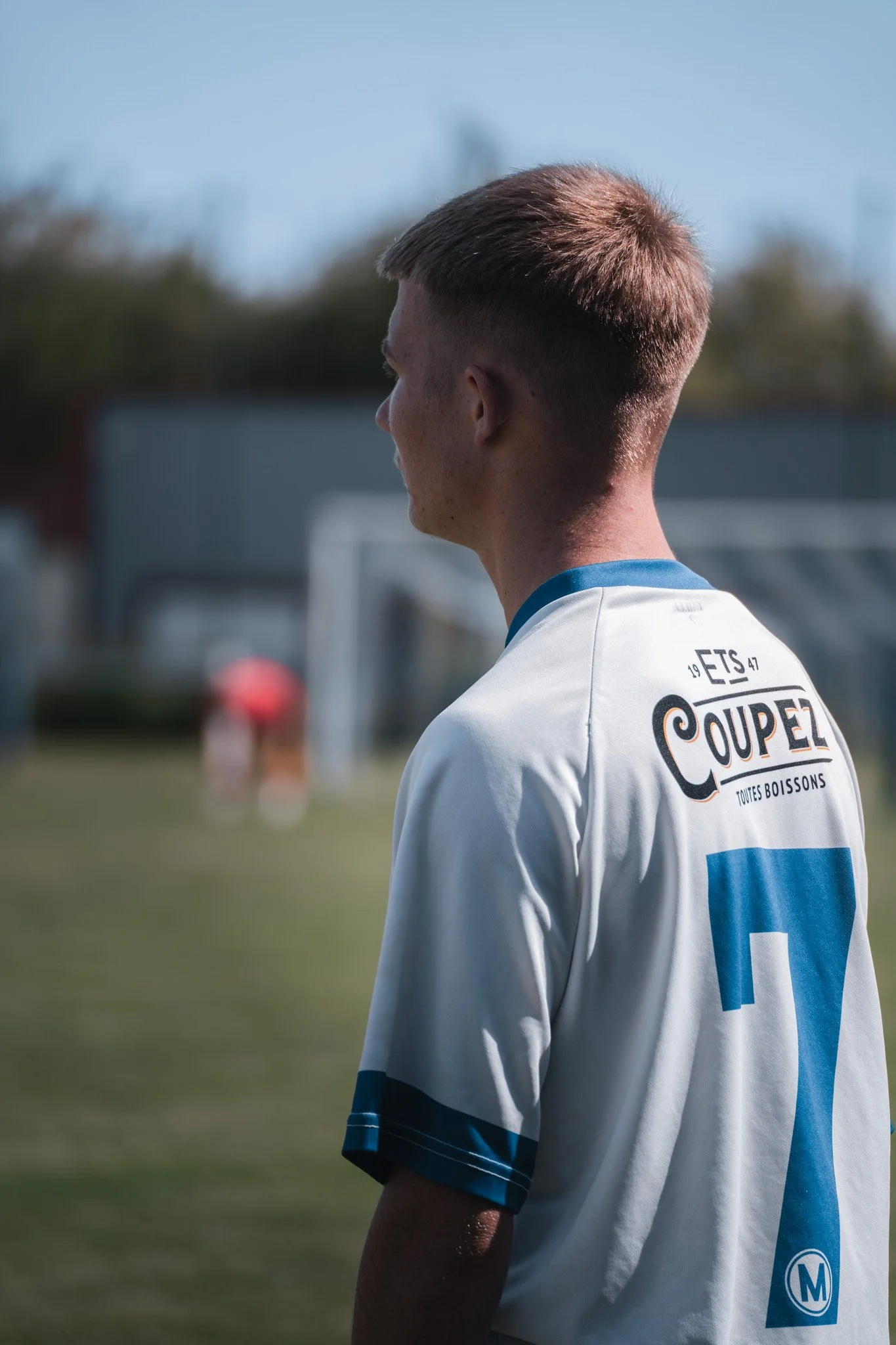 Jeune homme en maillot de football blanc et bleu, vu de profil, sur un terrain de football en plein air.