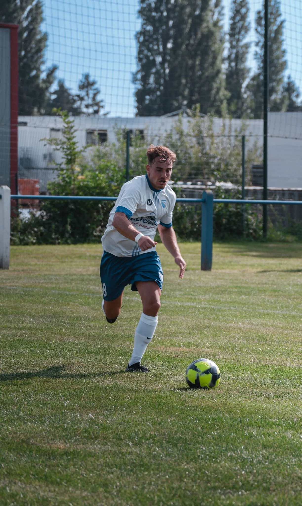 Joueur de football courant sur le terrain avec un ballon, portant un maillot blanc et un short bleu.