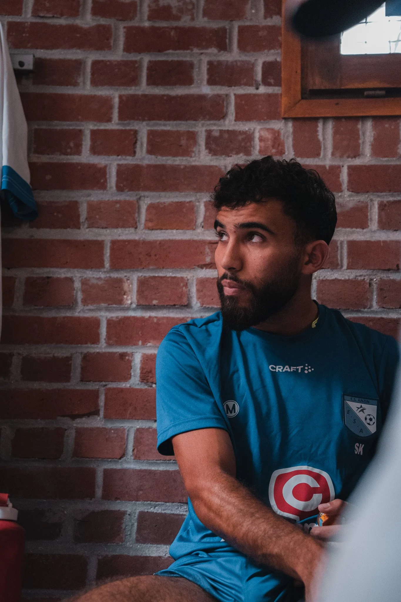 Un homme avec une barbe, portant un maillot de sport bleu, assis devant un mur en briques rouge, regardant sur le côté.