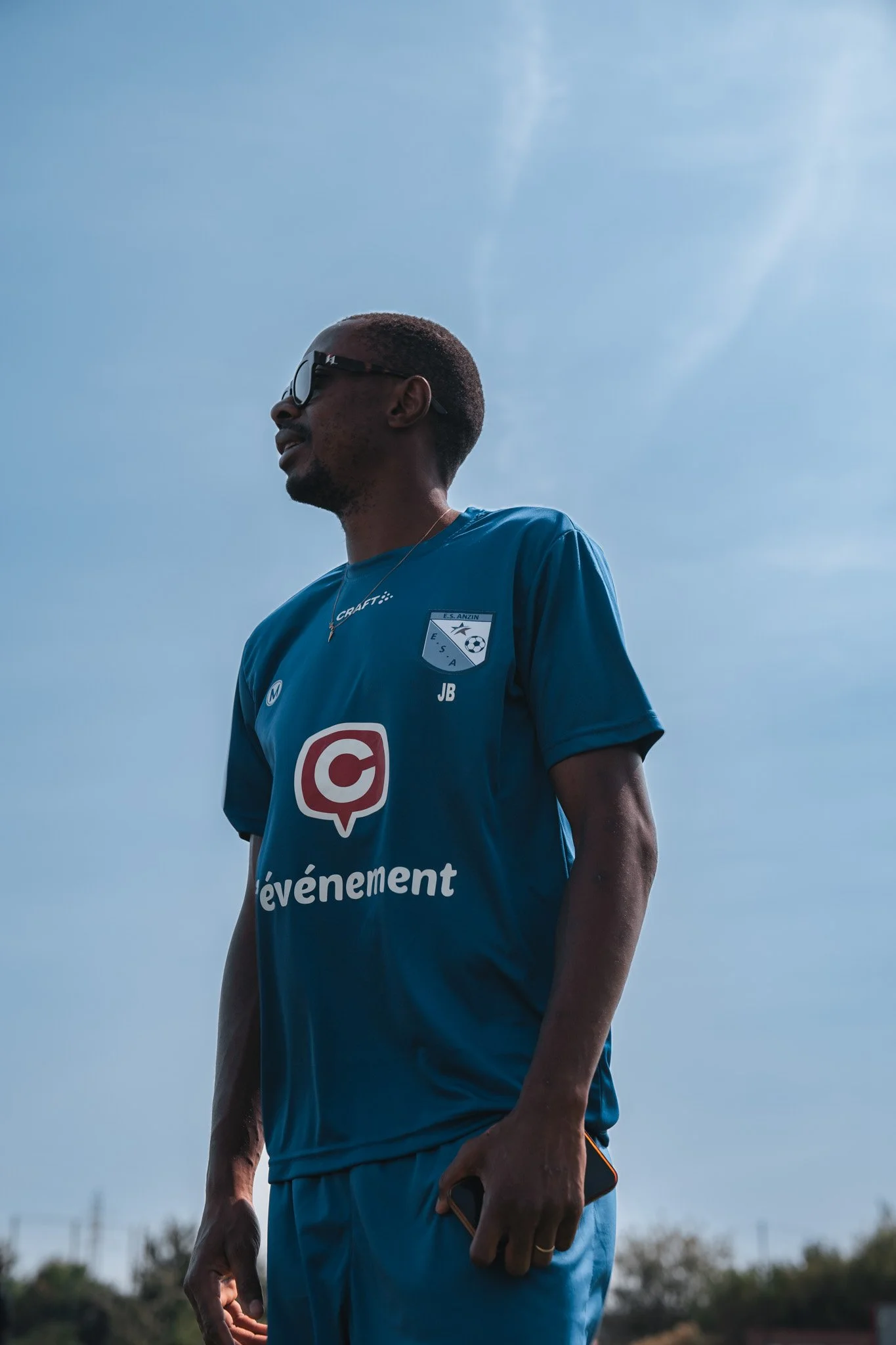 Un homme en maillot de sport bleu portant des lunettes de soleil, tenant un téléphone, dans un ciel bleu clair.