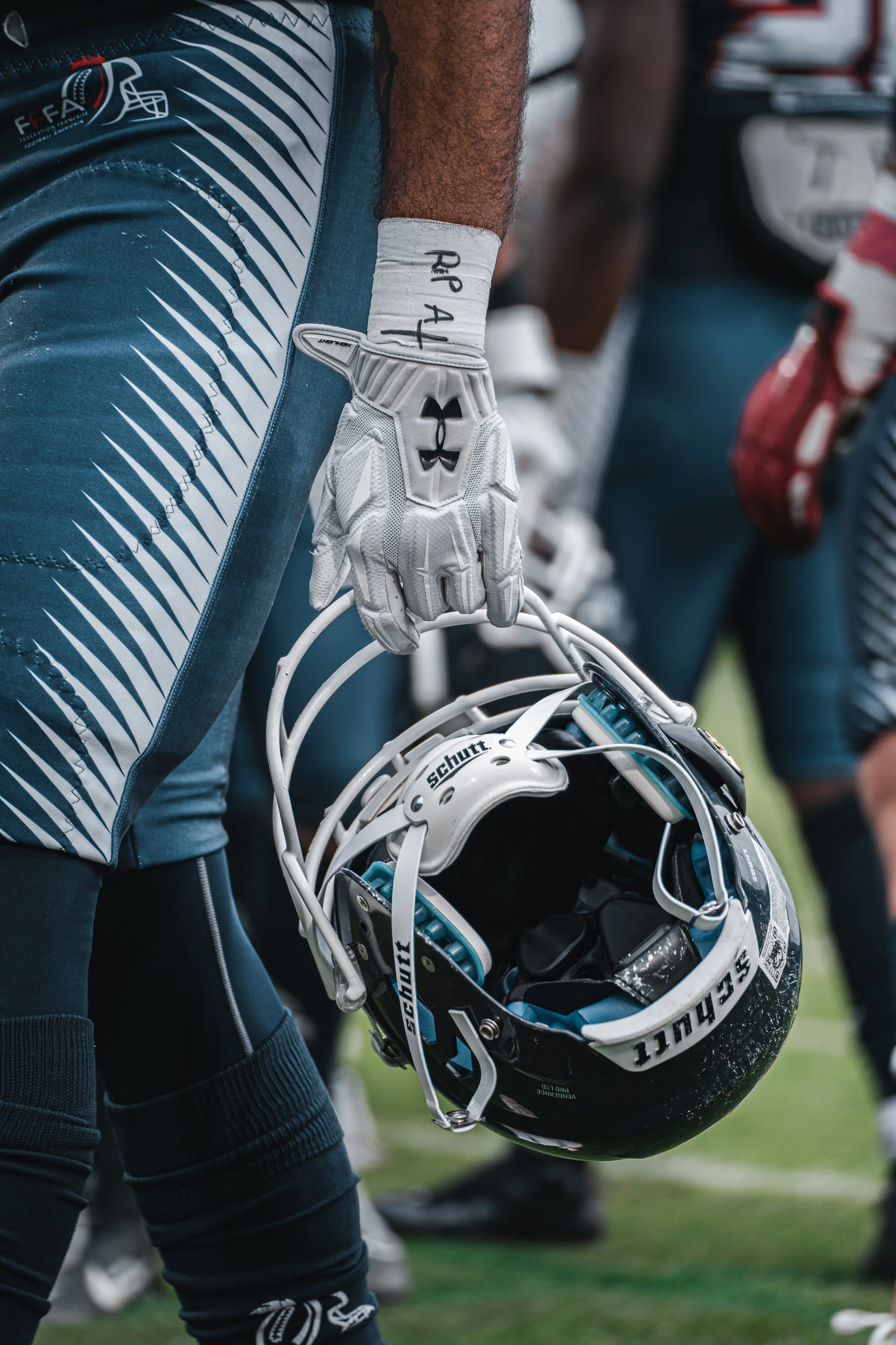 Un joueur de football américain tient un casque de football dans sa main gauche, portant un gant blanc et un uniforme avec des motifs bleus et blancs, en position sur le terrain lors d'un match ou entraînement.