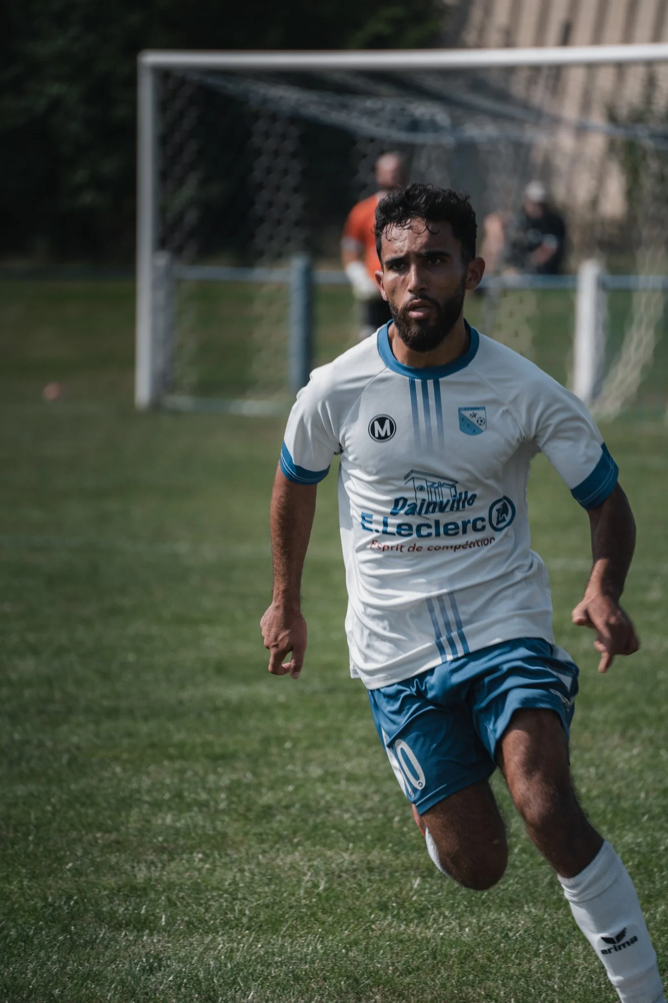 Un joueur de football court sur un terrain, portant un maillot blanc et bleu, avec un but en arrière-plan.