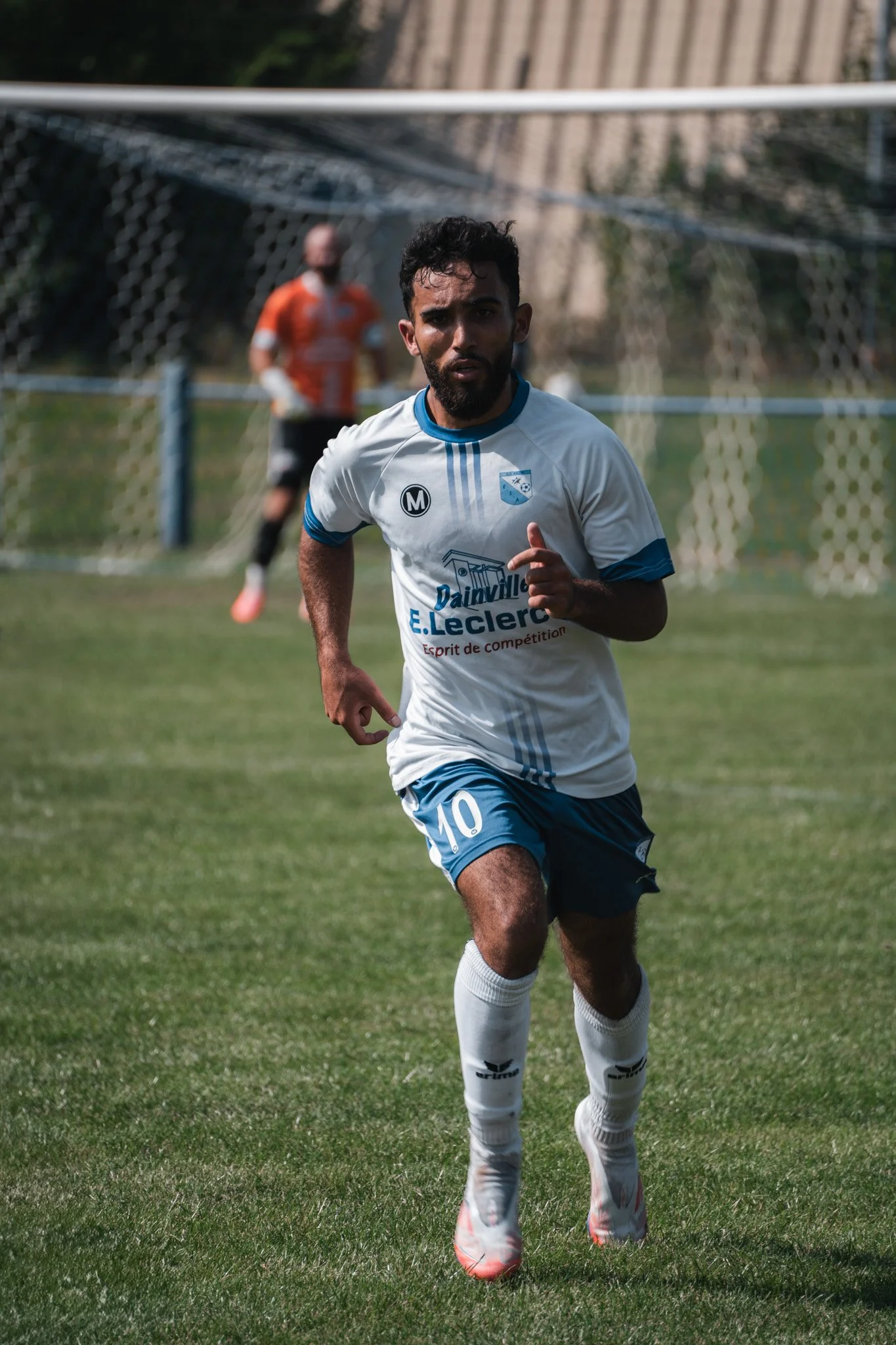 Un joueur de football en mouvement pendant un match, portant un maillot blanc avec le numéro 10, sur un terrain en herbe. En arrière-plan, un autre joueur en maillot orange.