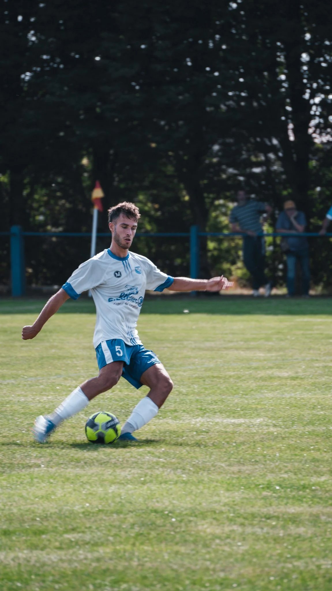 Jeune homme jouant au football sur un terrain en herbe, portant un maillot blanc et bleu, en train de préparer un coup de pied avec un ballon vert et noir.