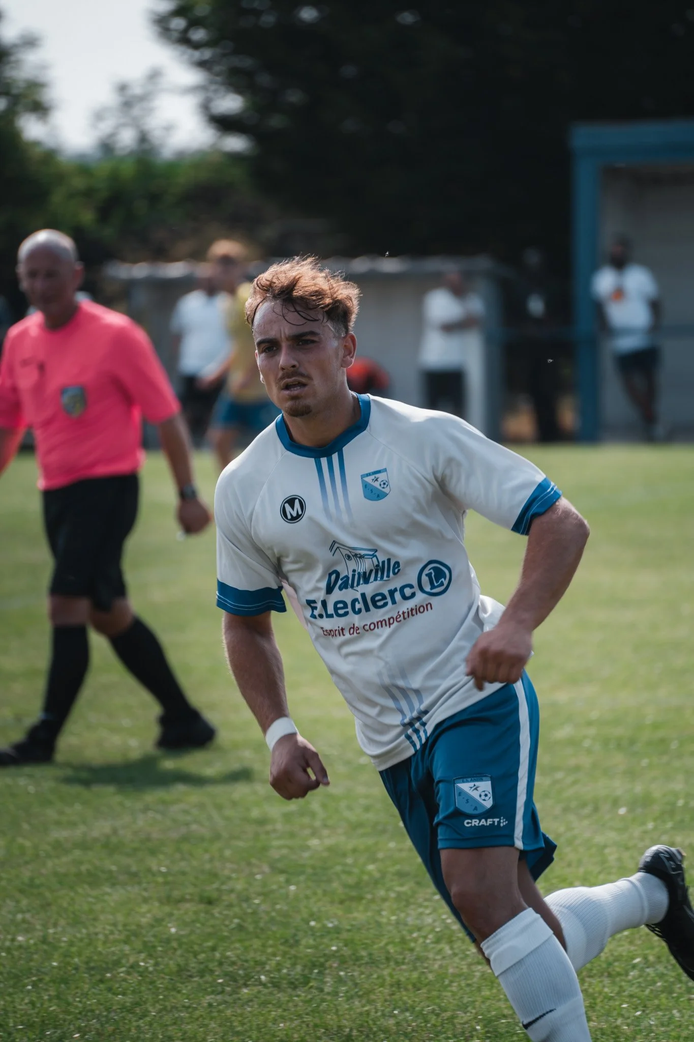 Un joueur de football en maillot blanc et bleu, en train de courir sur le terrain lors d'un match, avec un arbitre en rose en arrière-plan et quelques personnes observant depuis le bord du terrain.