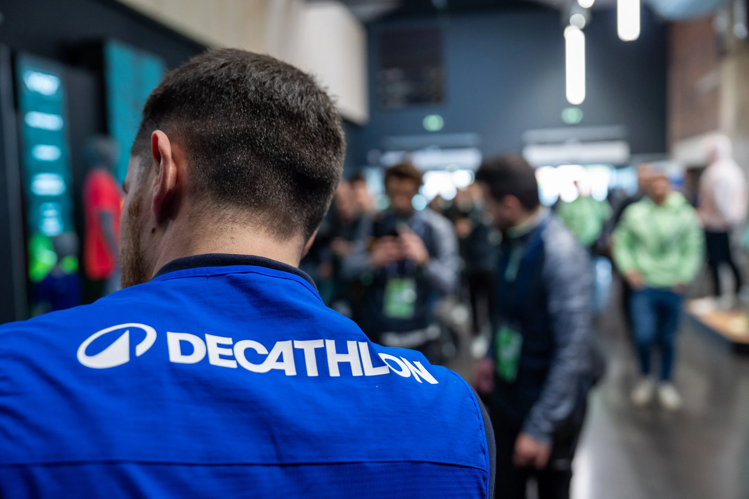 Un homme portant une veste bleue avec le logo 'DECATHLON' vu de dos, dans un environnement intérieur devant un groupe de personnes.