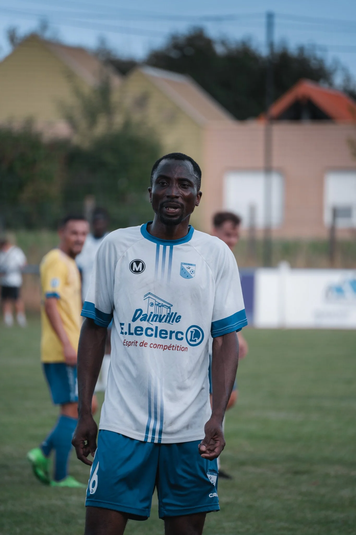 Un joueur de football en maillot blanc et bleu sur un terrain, avec d'autres joueurs en arrière-plan.
