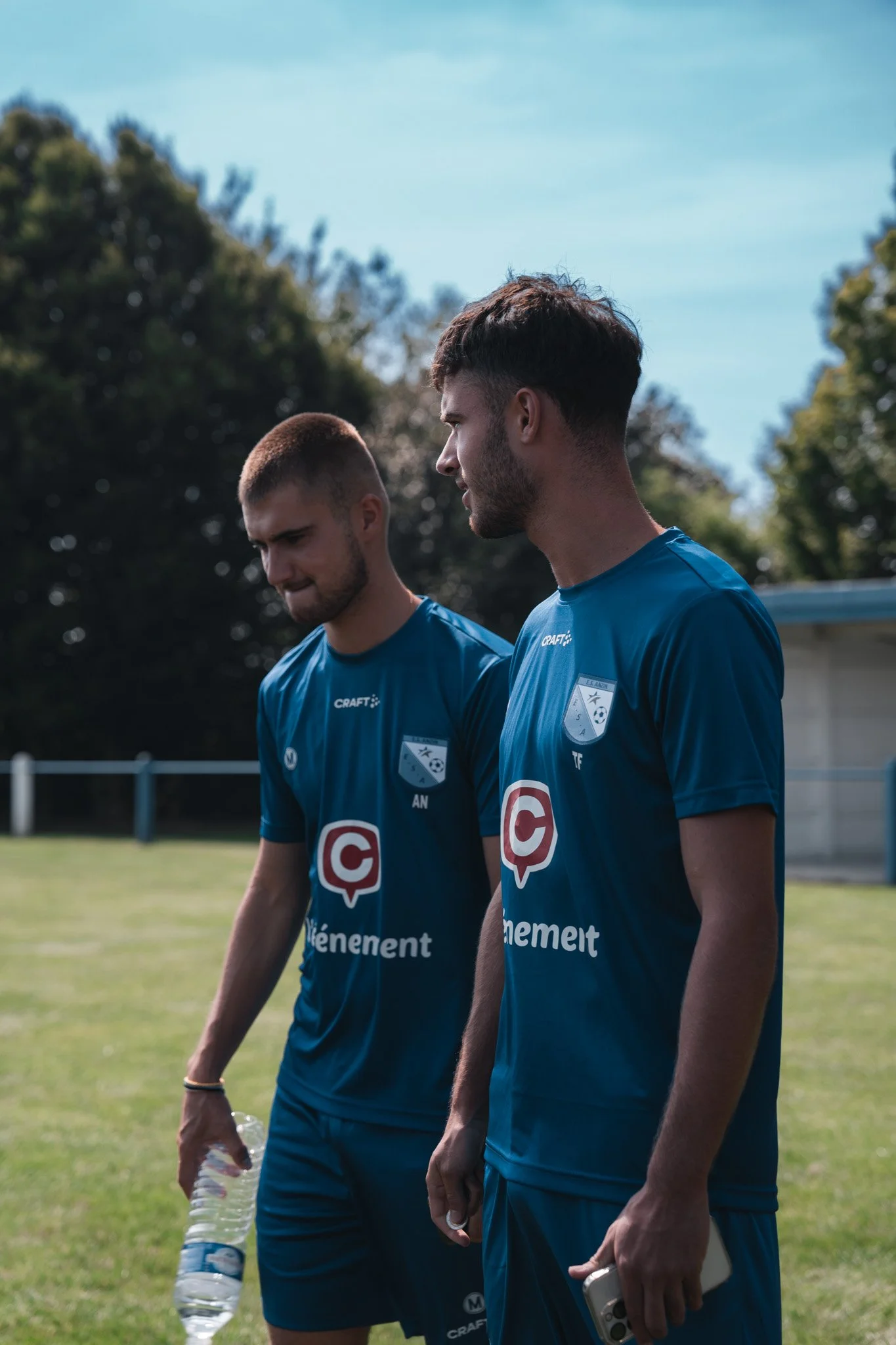 Deux jeunes hommes en maillots de football bleus sur un terrain en plein air, l'un tiendra une bouteille d'eau, l'autre un téléphone, avec des arbres en arrière-plan.