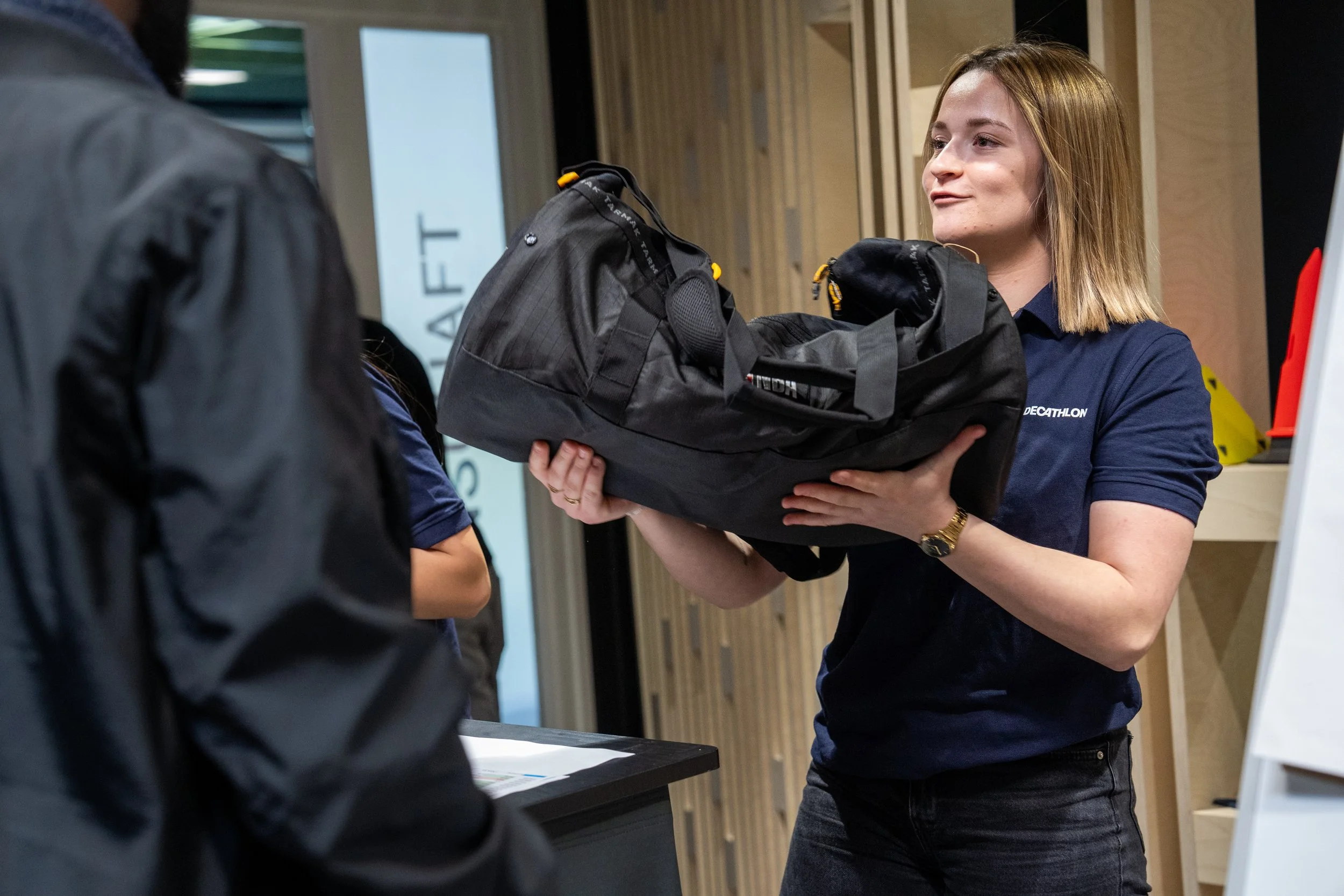 Une femme portant un t-shirt bleu avec le logo Decathlon, tenant un sac noir contenant des articles de sport, lors d'une interaction en intérieur.