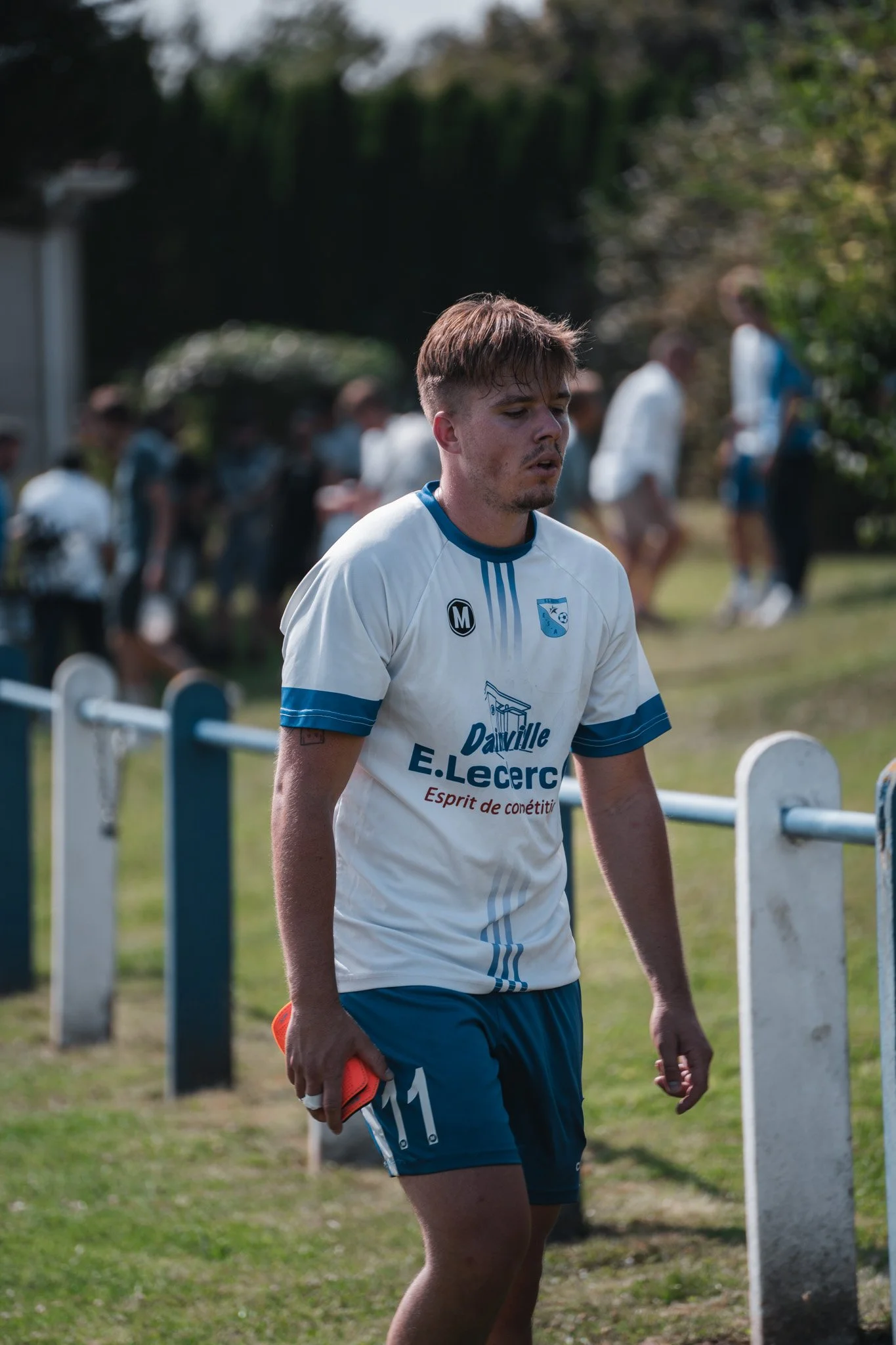 Un joueur de football en tenue blanche et bleue, portant le numéro 11, marchant sur le terrain avec un arrière-plan flou de spectateurs et de nature.