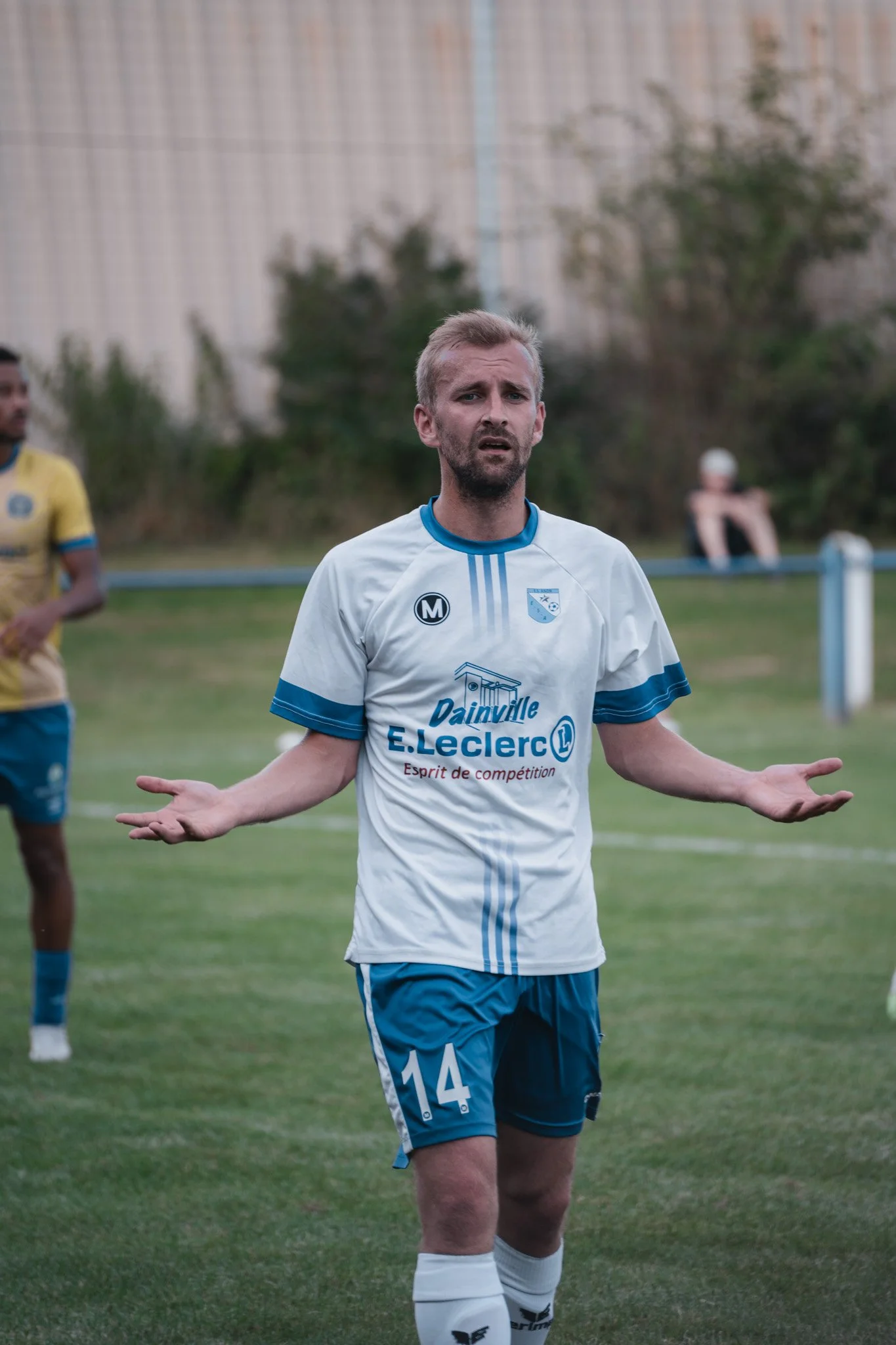 Un joueur de football avec un maillot blanc et bleu, portant le numéro 14, a une expression confuse ou déconcertée, sur un terrain de football.