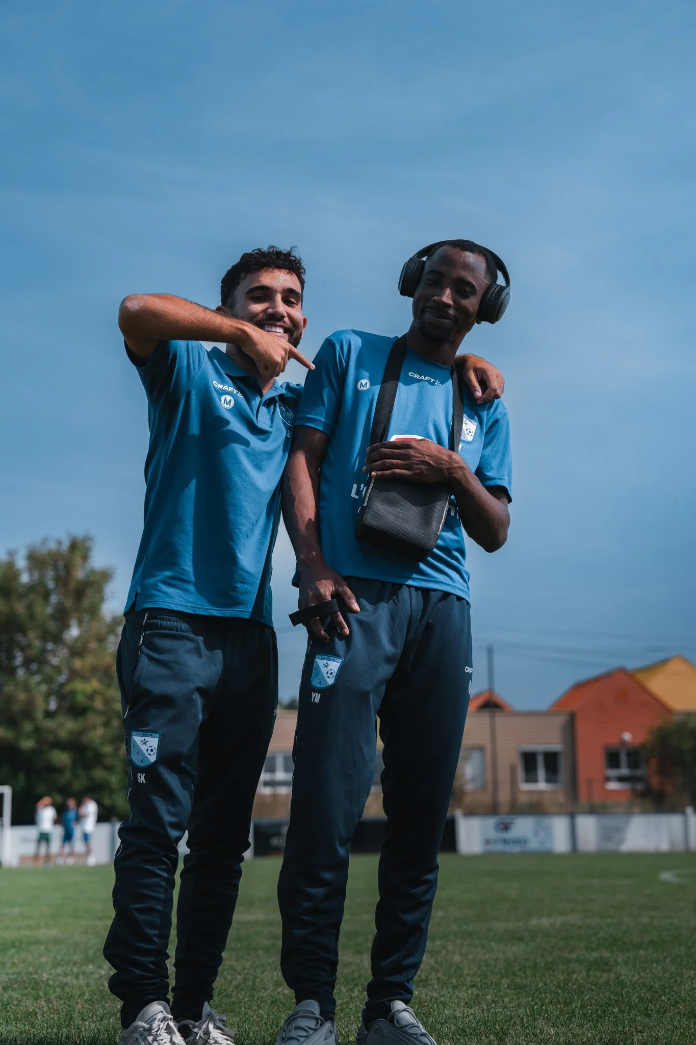 Deux hommes en tenues de sport bleues sur un terrain de sport, l'un avec un casque audio, souriants, en train de poser pour la photo.