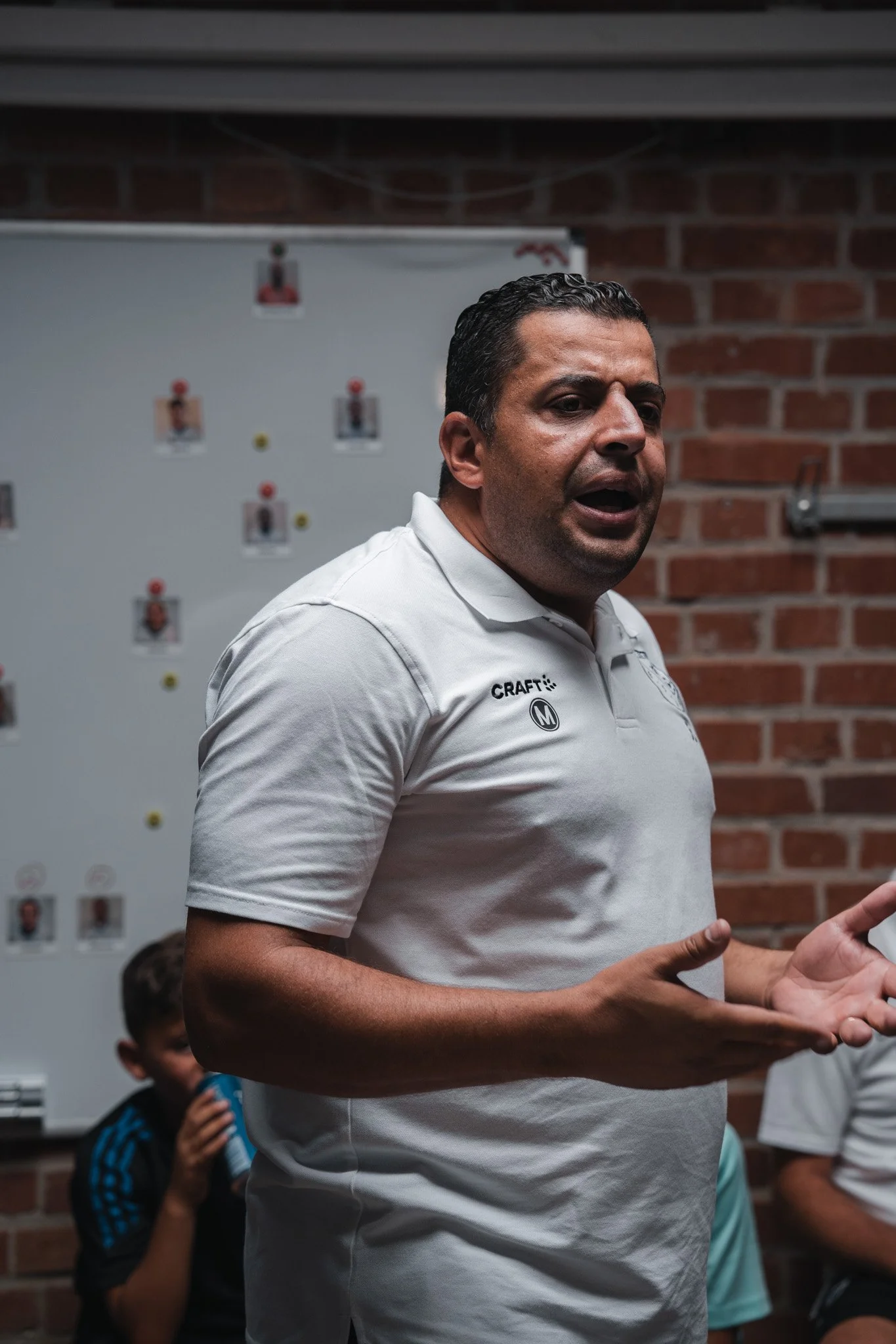 Homme en polo blanc parlant devant un groupe d'enfants dans une pièce avec mur en briques rouges et tableau blanc.