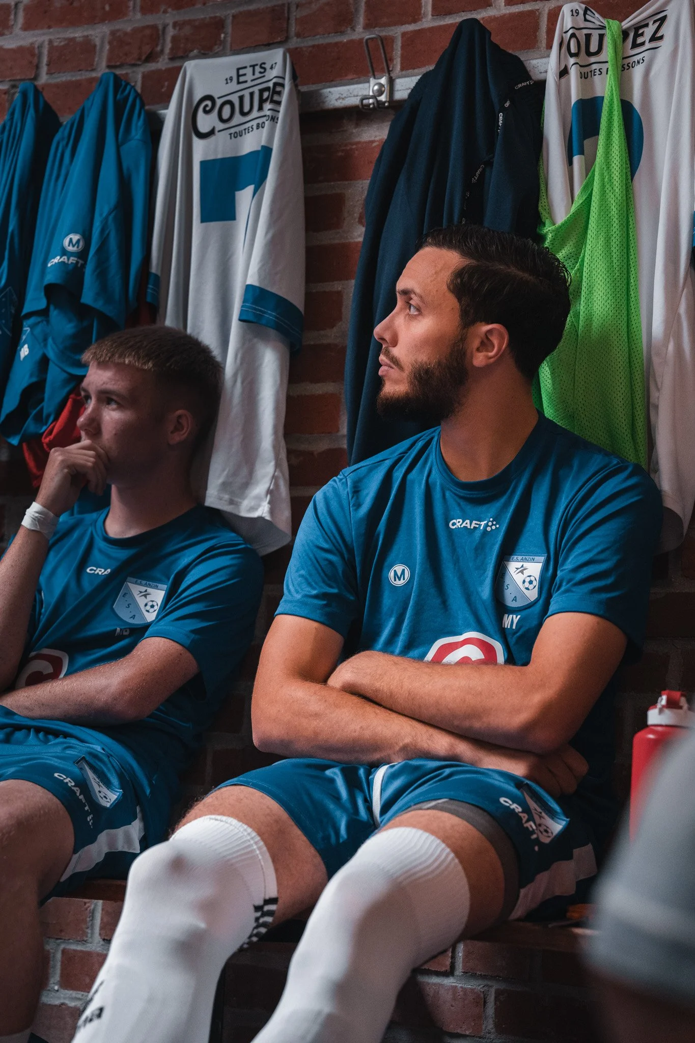Deux joueurs de football en tenue dans une salle de vestiaire, assis sur un banc, avec des maillots de football suspendus derrière eux sur un mur en briques rouges.