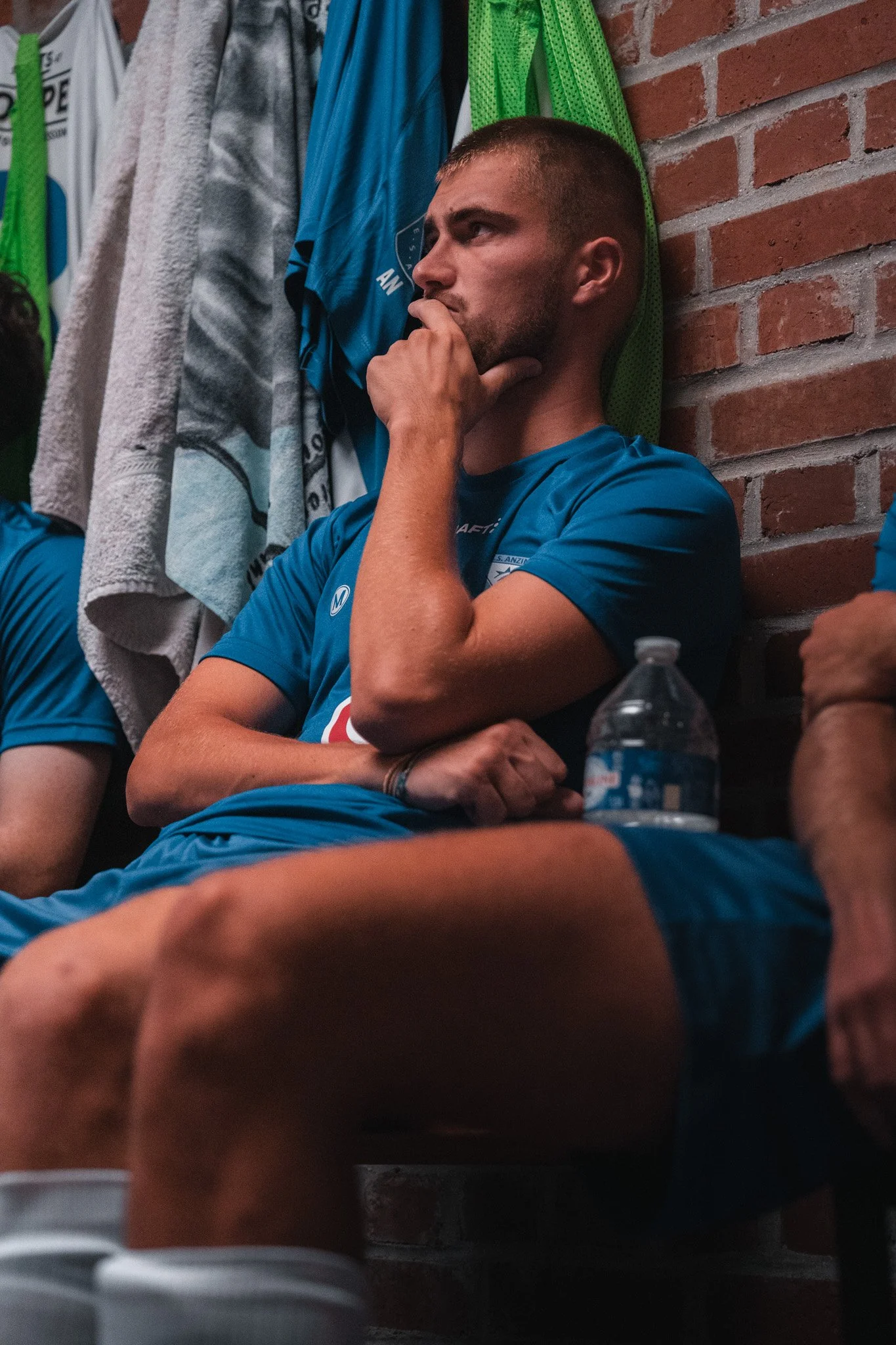 Un homme assis dans un vestiaire, portant un maillot de sport bleu, semble réfléchir ou être préoccupé, avec une bouteille d'eau à côté.
