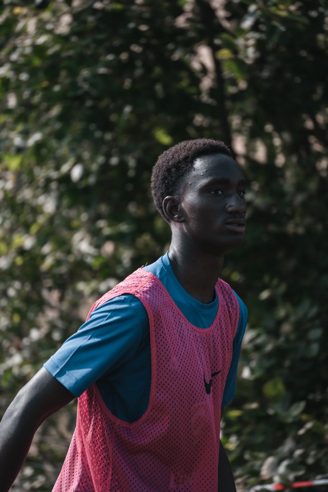 Jeune homme athlétique avec peau foncée, portant un maillot de sport bleu et un gilet rose, se tenant à l'extérieur devant un fond de feuillage vert.