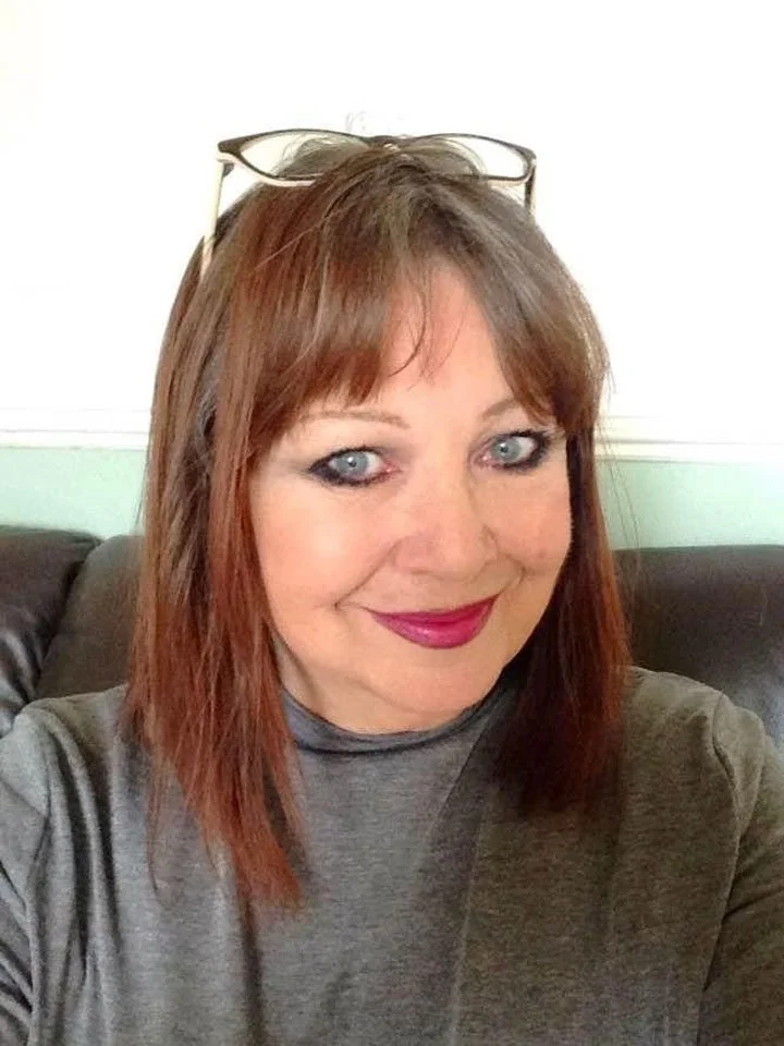 Jackie is a white woman with short red hair and a fringe. She is wearing a grey top and bright lipstick.