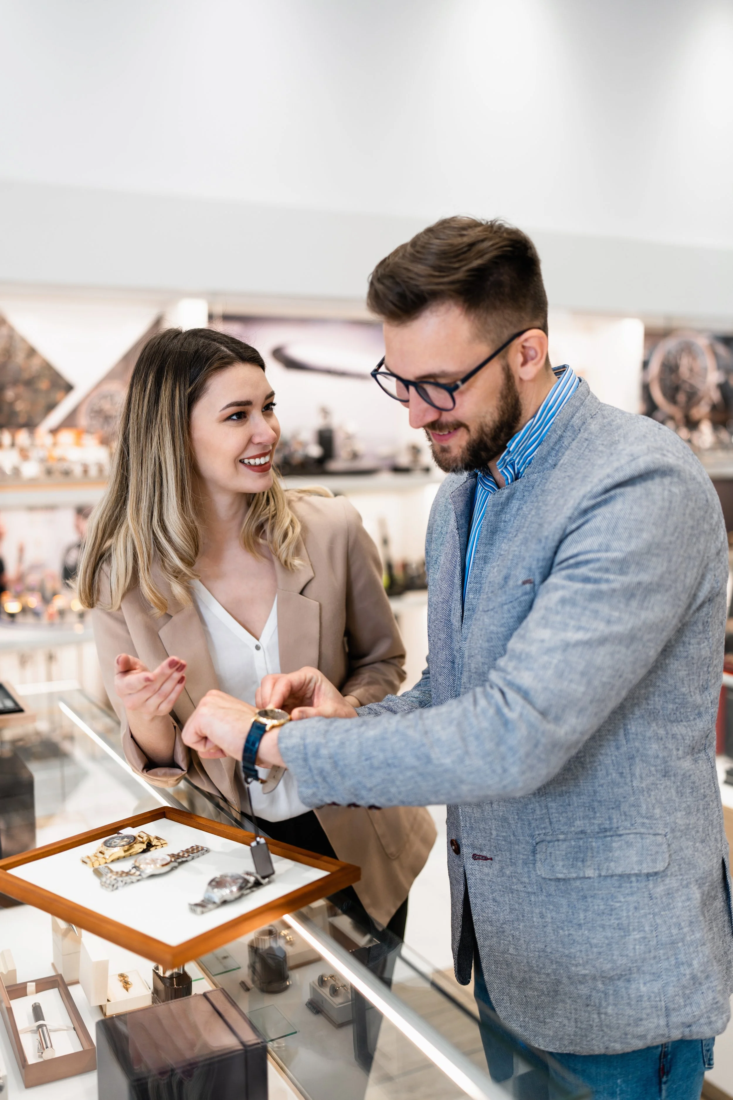 A man and woman shopping for watches at a jewelry store, with the man trying on a watch and the woman looking at him, on a glass display case filled with watches.