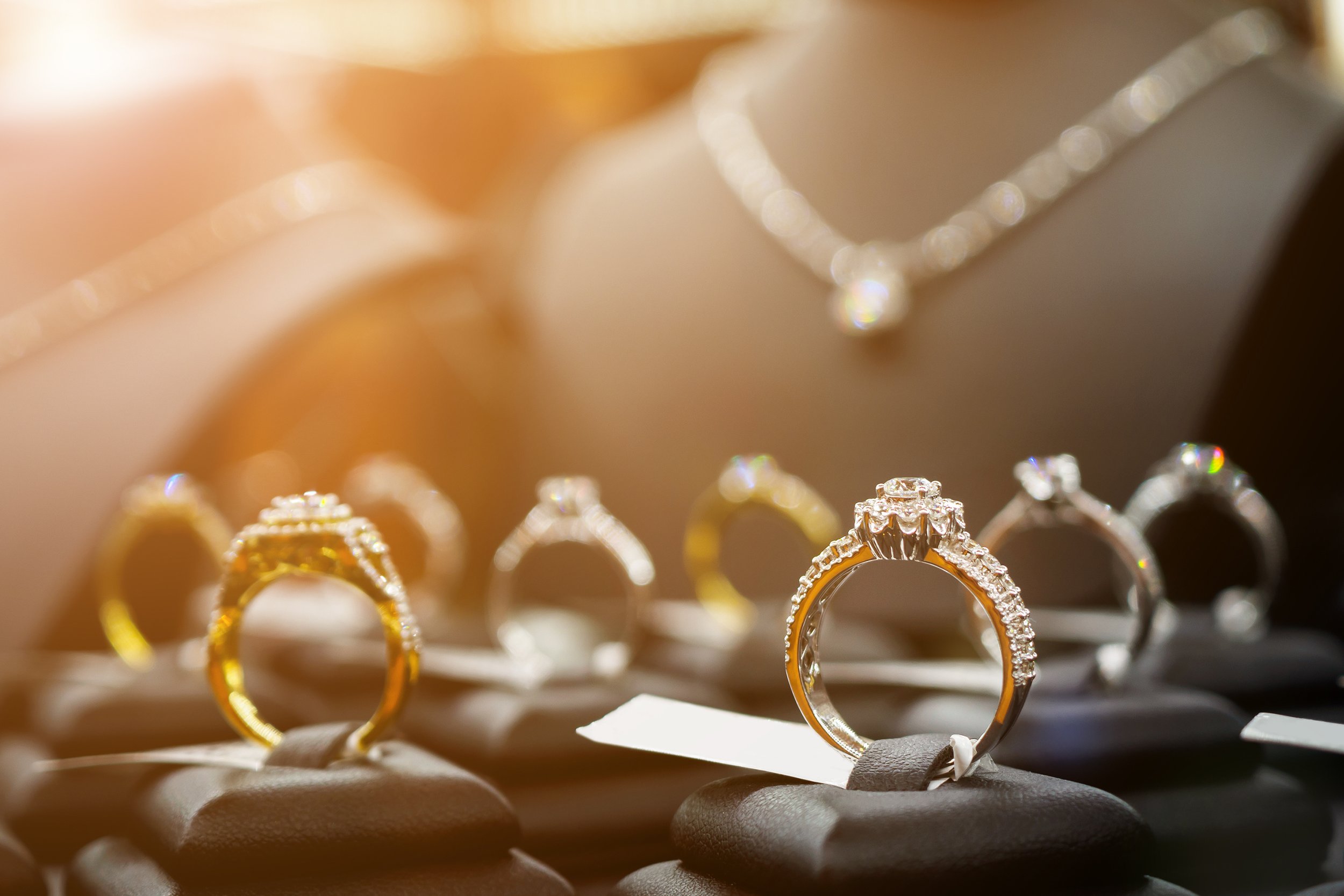 Close-up of various diamond engagement rings and wedding bands displayed on black ring holders, with a focus on the central ring with a large diamond, in soft warm lighting.