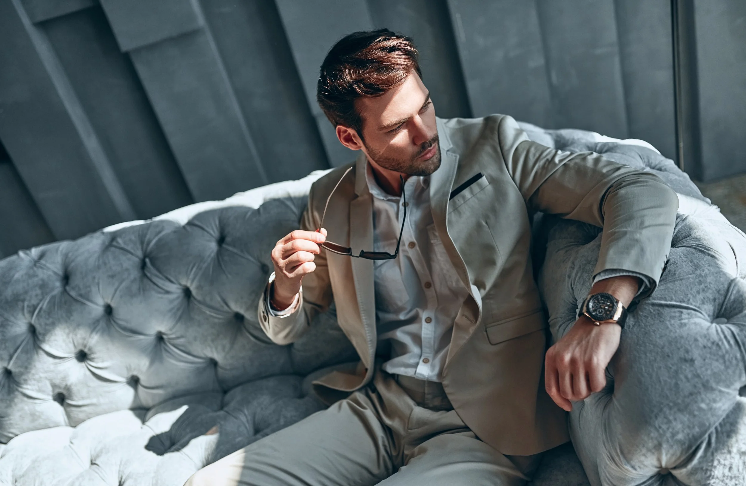 A man sitting on a light gray tufted sofa, holding sunglasses in his right hand, wearing a beige suit, white shirt, wristwatch, and looking down thoughtfully.