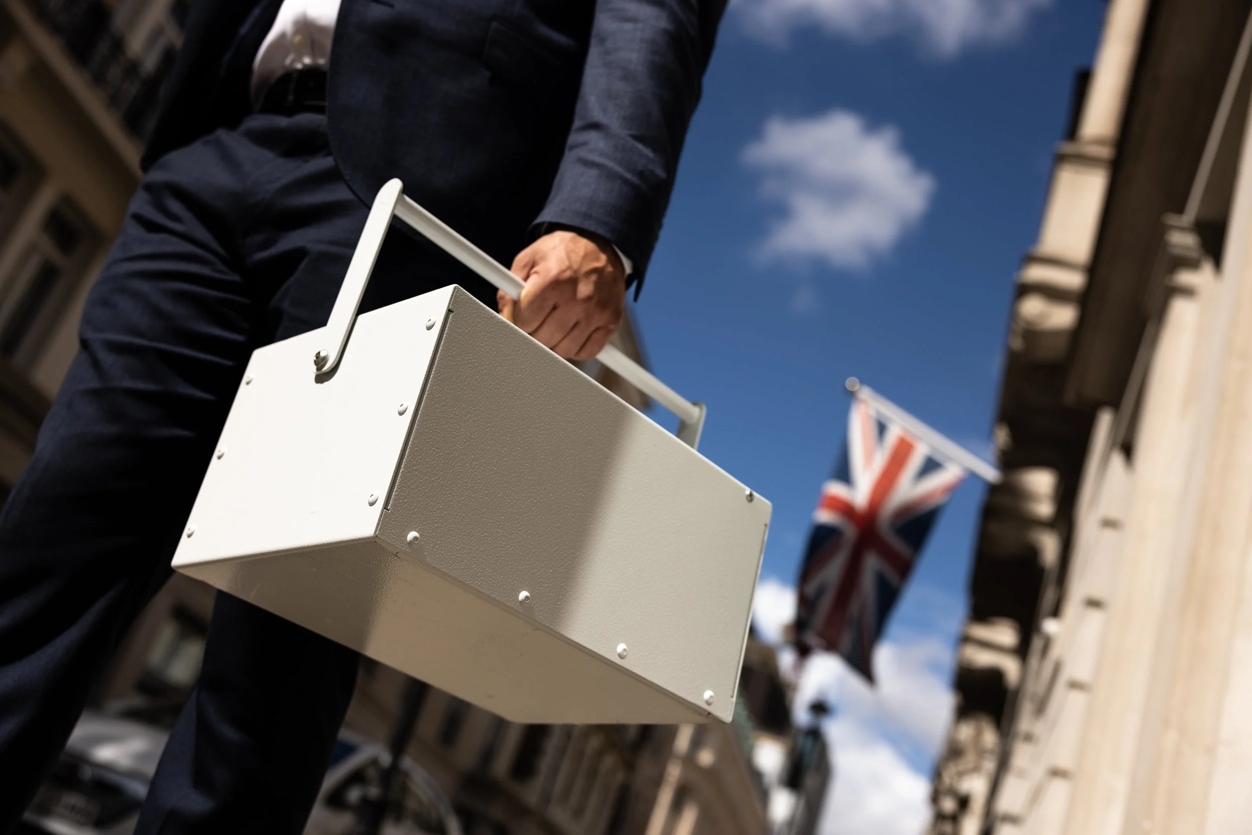 Close-up of a person in a dark suit holding a beige briefcase on a city street with Union Jack flags and historic buildings in the background.
