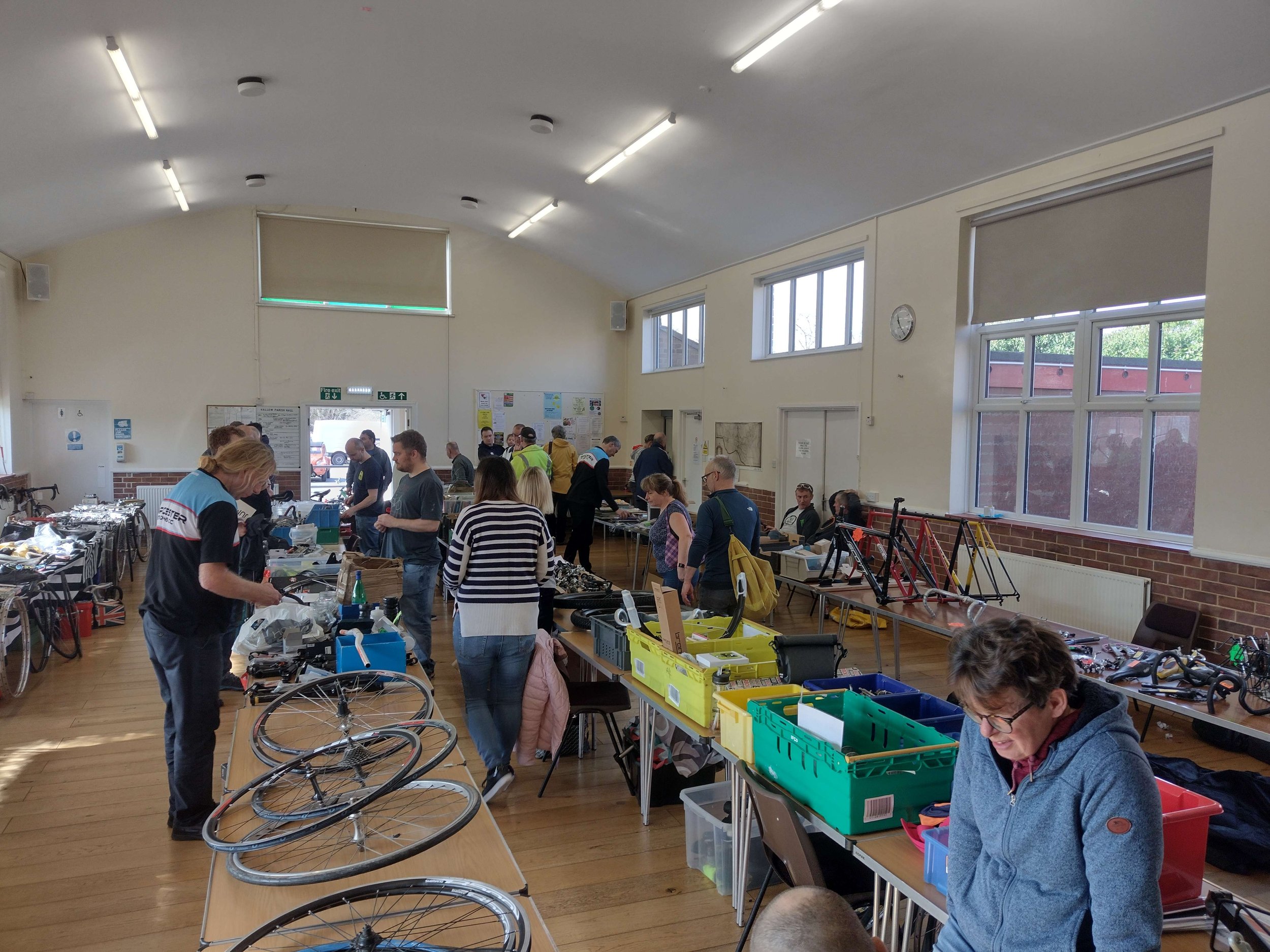 Boxes of bicycle parts, frames and wheels are laid out on tables in a large village hall with people browsing the items.