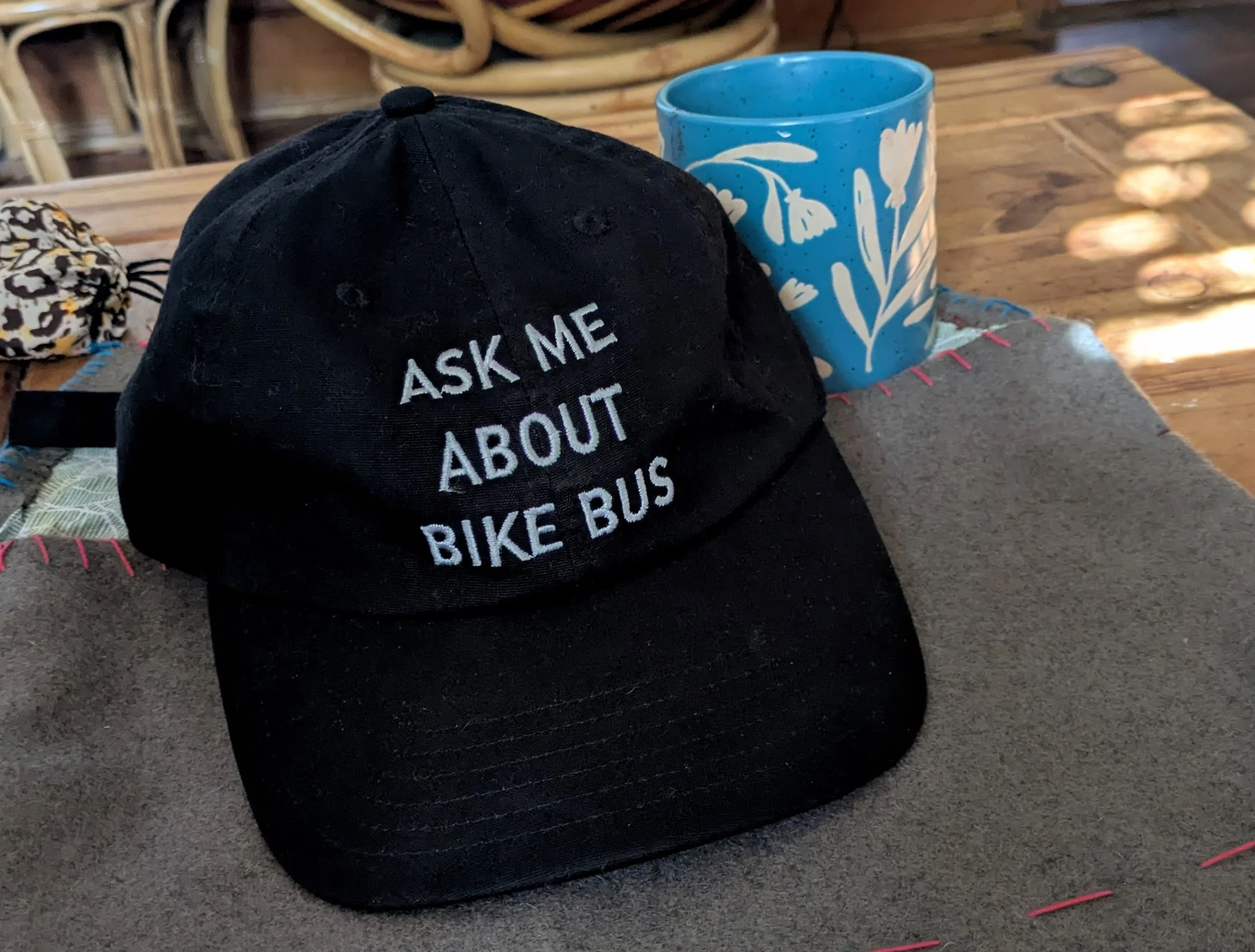 A black baseball cap next to a cup. The hat has embroidery that says "Ask me about Bike Bus".