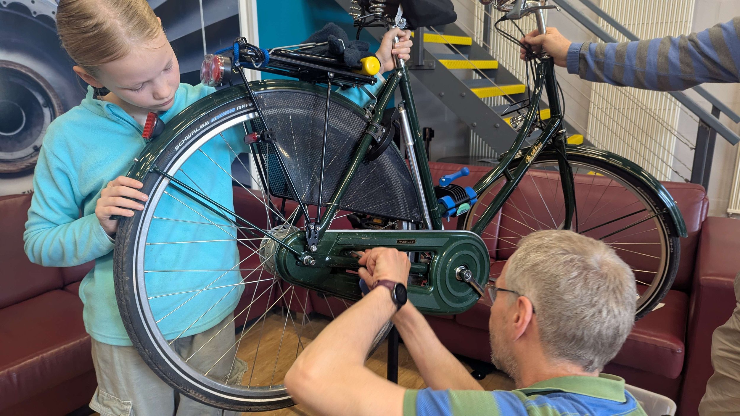 A bicycle mounted in a stand is repaired by a volunteer and their daughter.