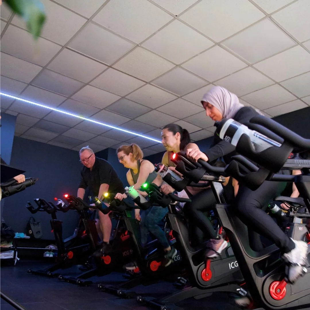 Group of people participating in a spinning class on stationary bikes in a gym, illuminated by colorful LED lights.