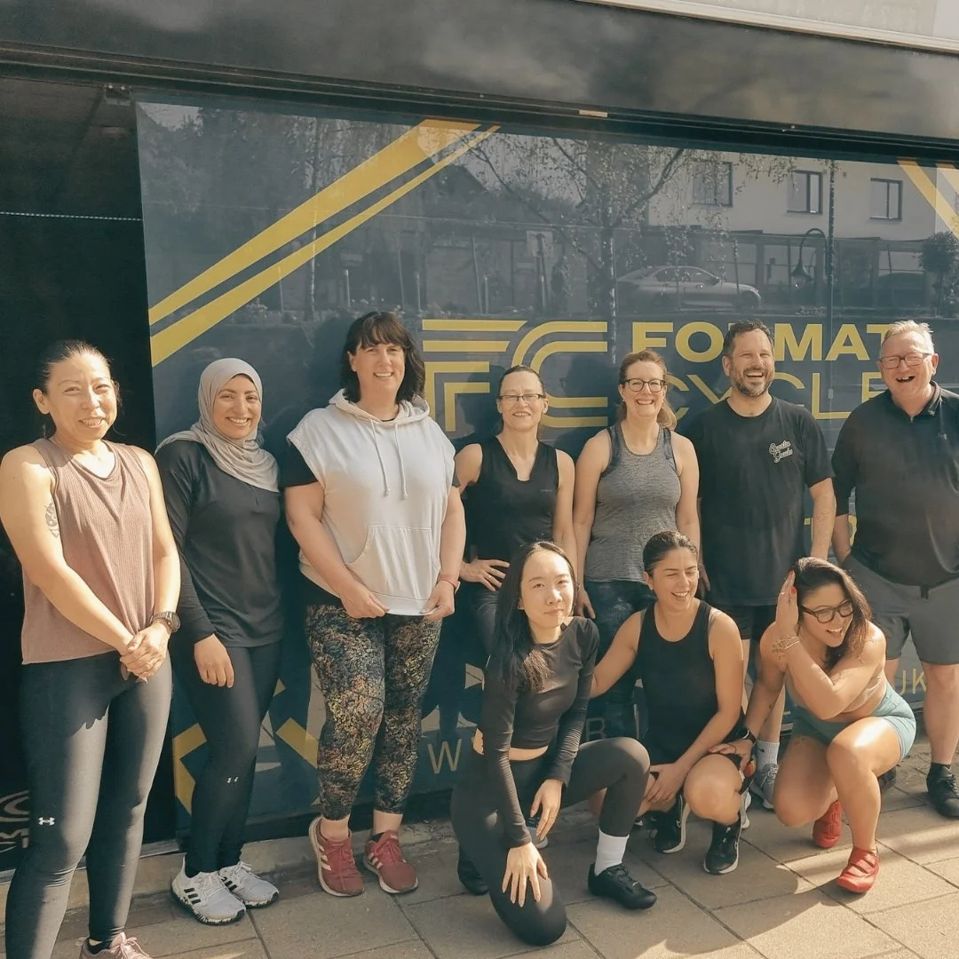 Group of ten people, mostly women, smiling and posing for a photo outside in front of a black and yellow sign that says 'Format' or 'Formula'. Some are standing, some kneeling, dressed in casual athletic wear.
