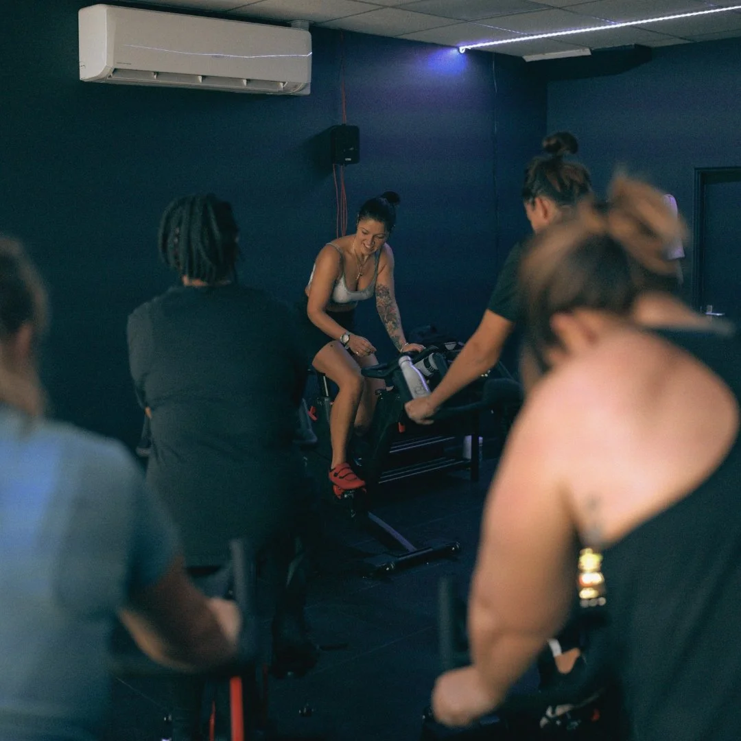 A group of women participating in an indoor fitness class, with one woman on a stationary bike in focus, smiling and engaging with the instructor, in a dimly lit exercise room.