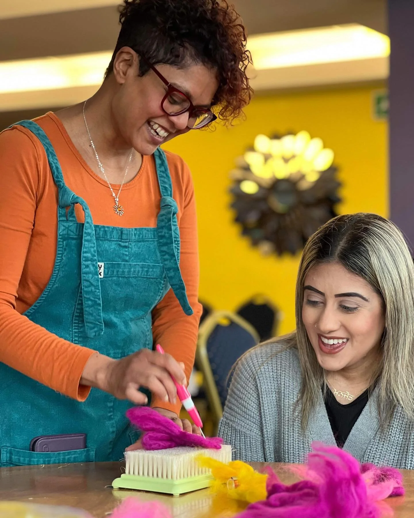 Two women looking at colorful yarn and a brush on a table, smiling and engaging in a craft activity in a brightly lit room.