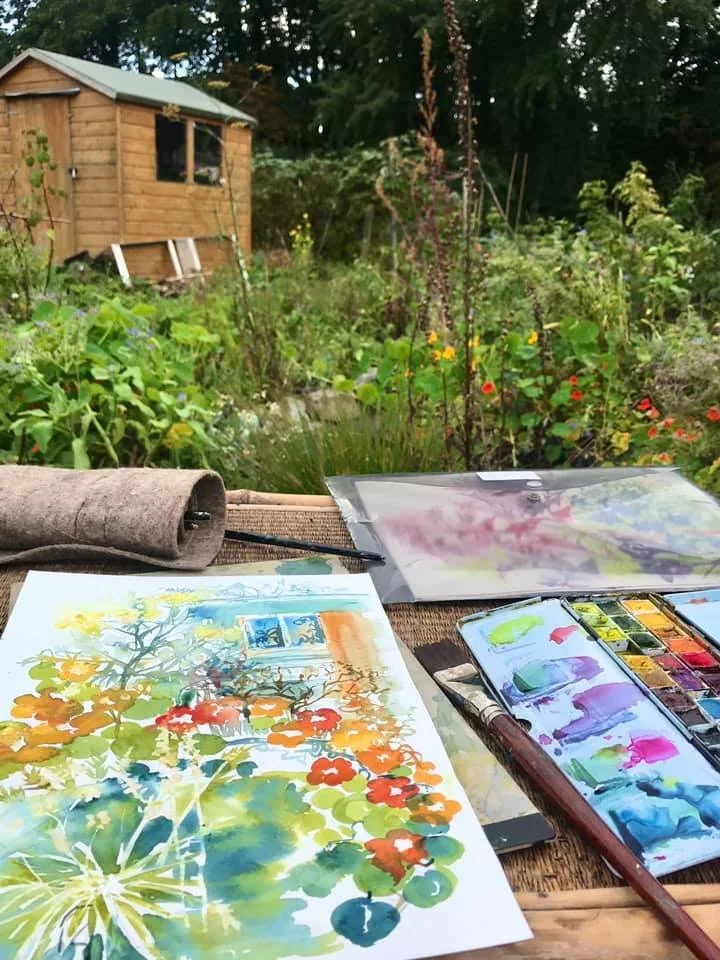 Watercolor painting supplies on a wooden table outdoors, with a garden and a wooden shed in the background.