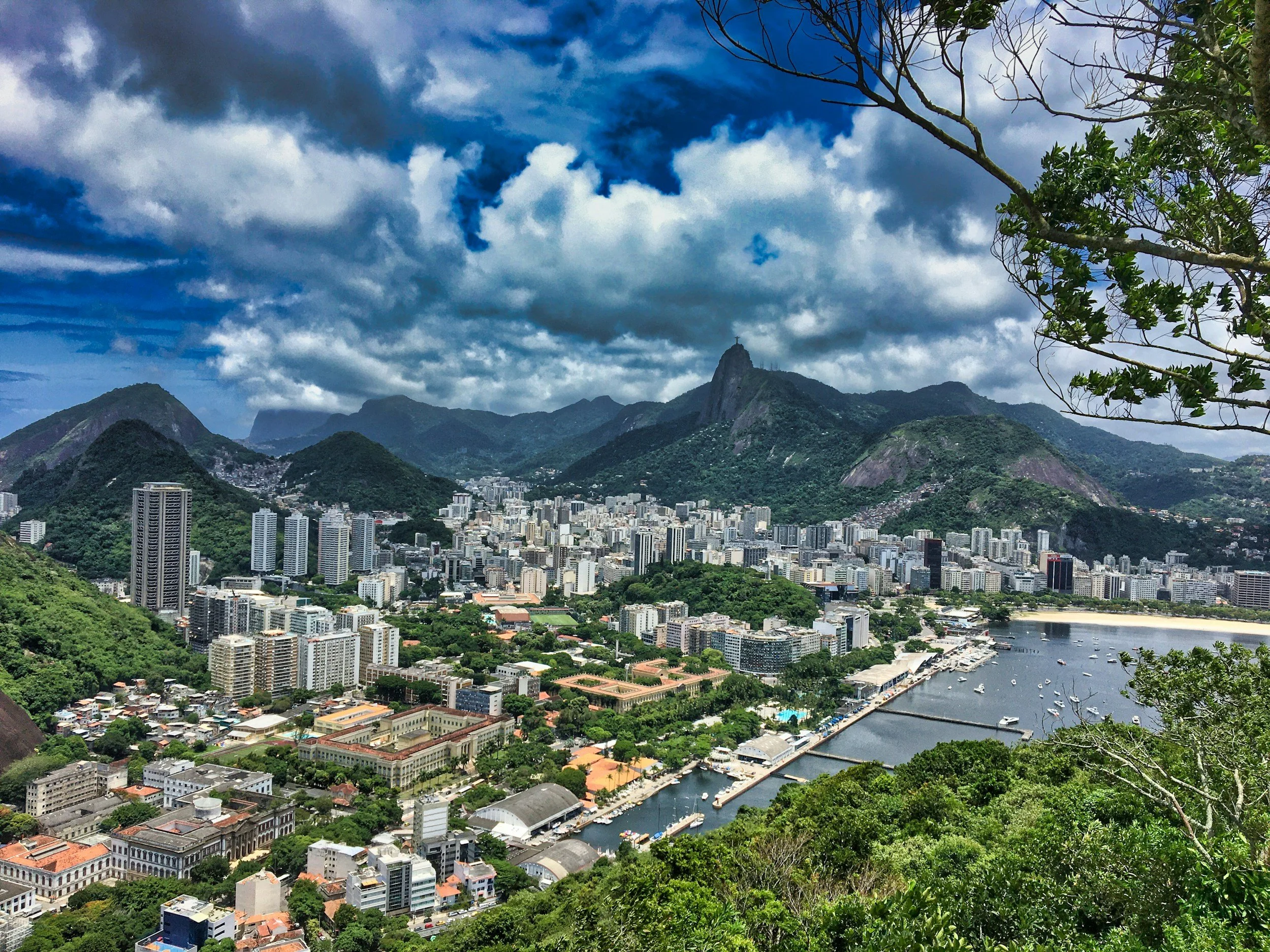Aerial view of Rio de Janeiro showing mountains, city buildings, and a harbor with boats, under a partly cloudy sky.