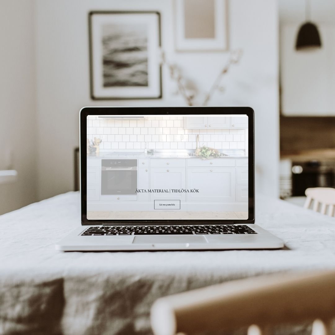 Hemsida för Stockshammar kök och inredningsdesign uppsatt av Isabel Azcárate Din Marketing Coach och Johanna Egemar, företagsfotograf. En öppen laptop på ett vitt bord i ett kök med vit inredning och bilder på väggen i bakgrunden.