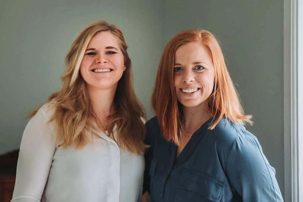 Två kvinnor som ler, Isabel Azcárate Din Marketing Coach och Johanna Egemar, företagsfotograf, står nära ett fönster mot en grön vägg.