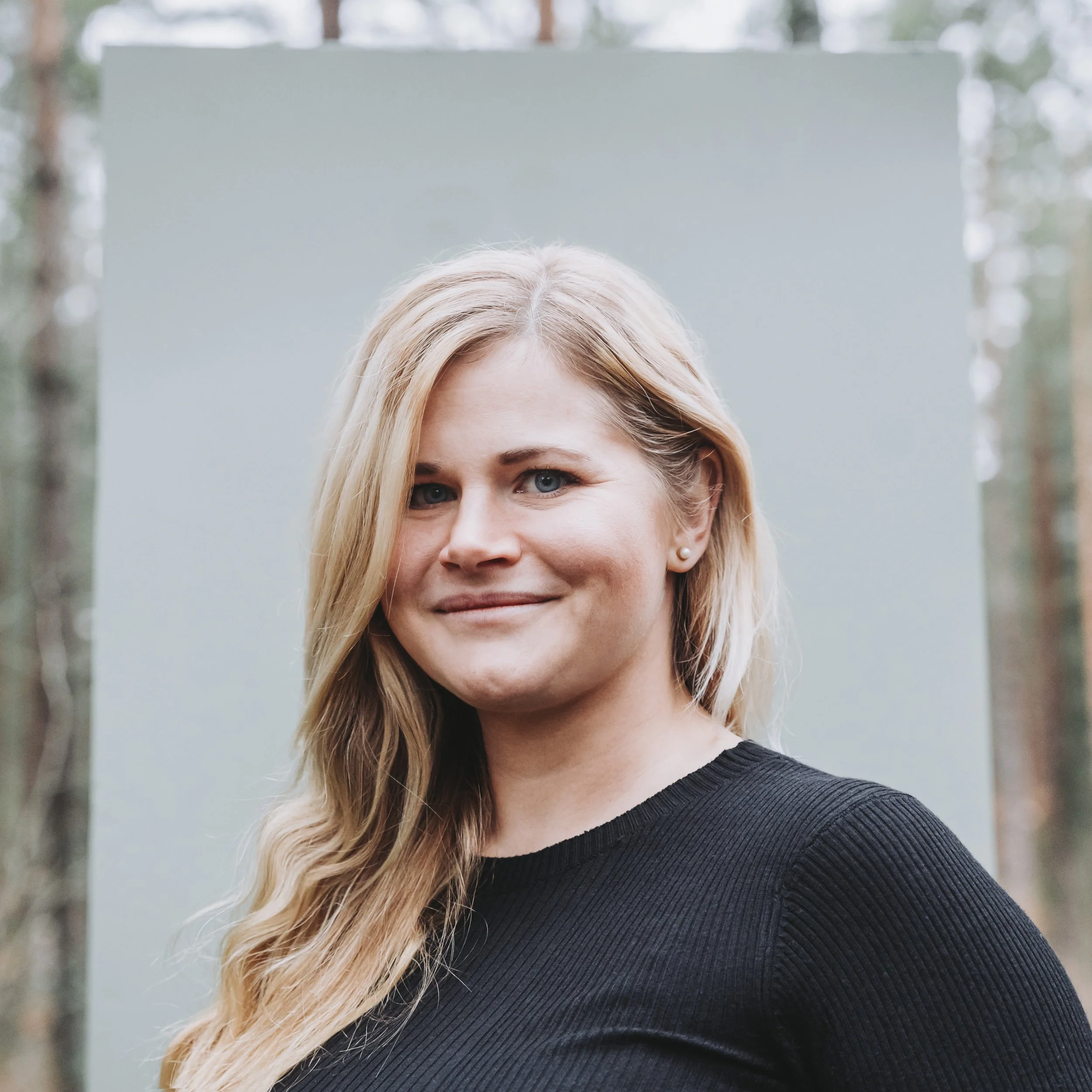 En kvinna med långt blont hår och blå ögon, Isabel Azcárate Din Marketing Coach klädd i en svart tröja, ler lätt, står utomhus.