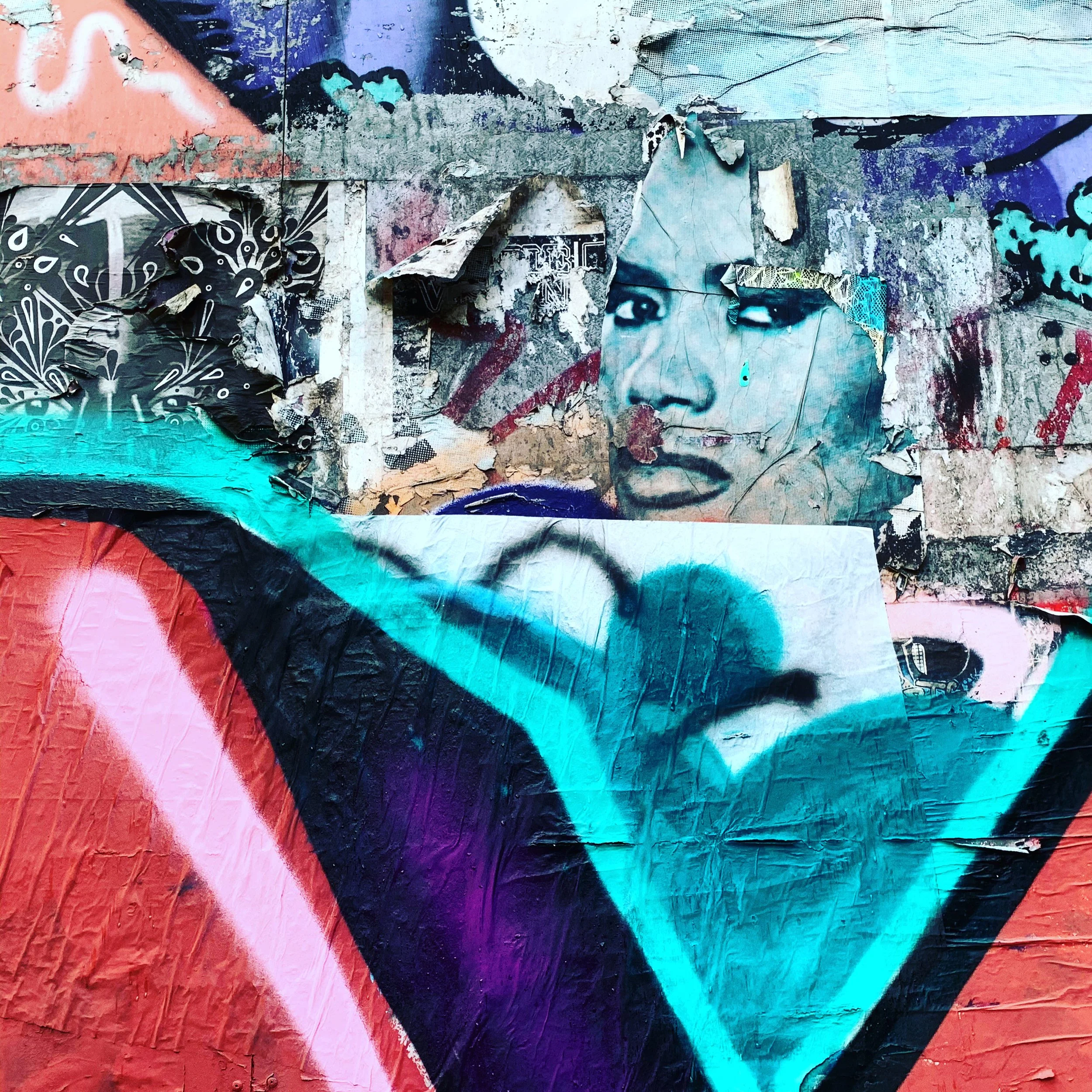 Street art mural featuring a woman's face with makeup and styled hair, surrounded by colorful graffiti and posters with layers of torn paper.