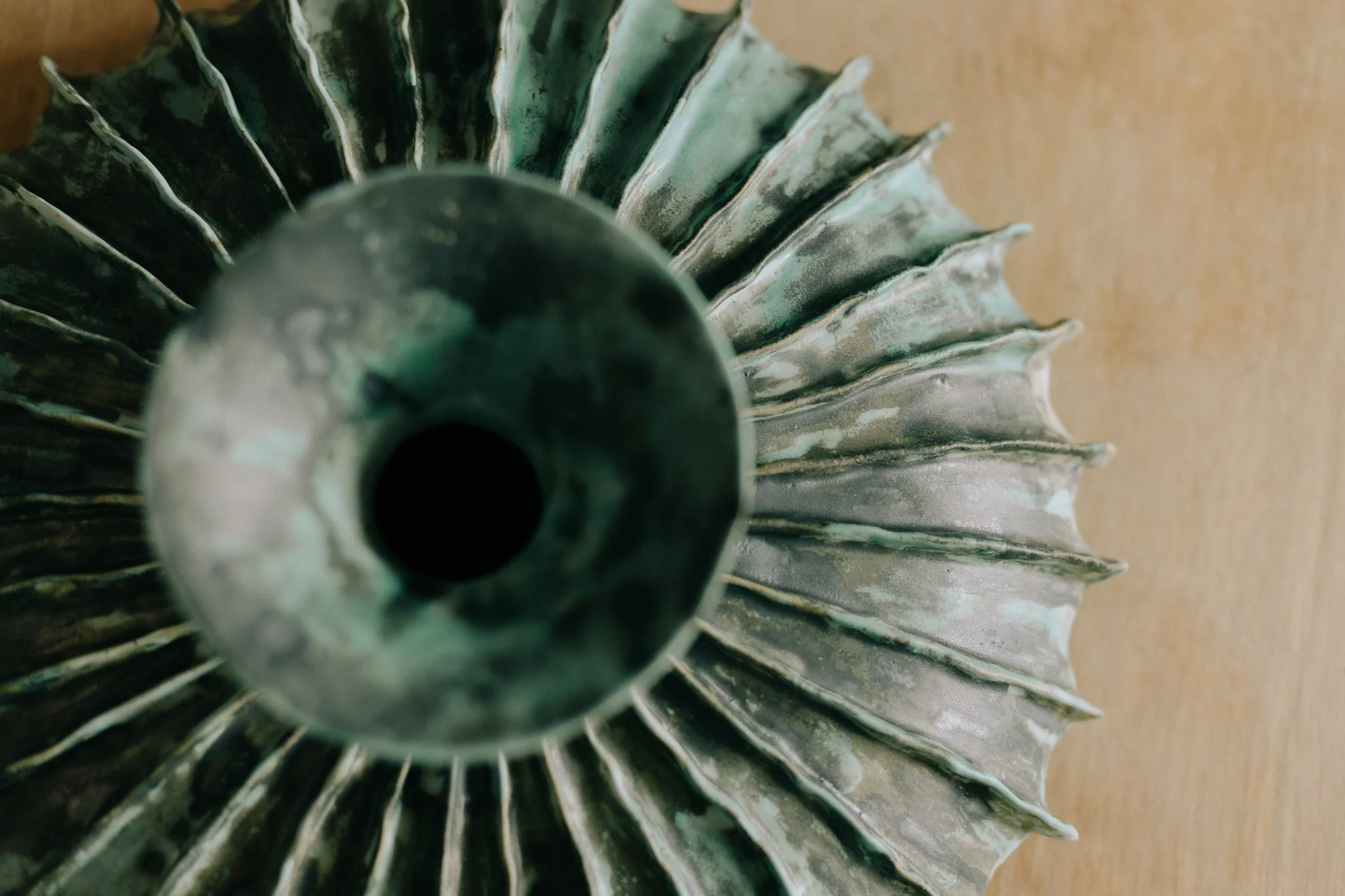 Close-up view of an antique decorative metal object resembling a sunburst or funnel with pointed ridges radiating outward, with a circular opening at the center, placed on a light brown textured surface.