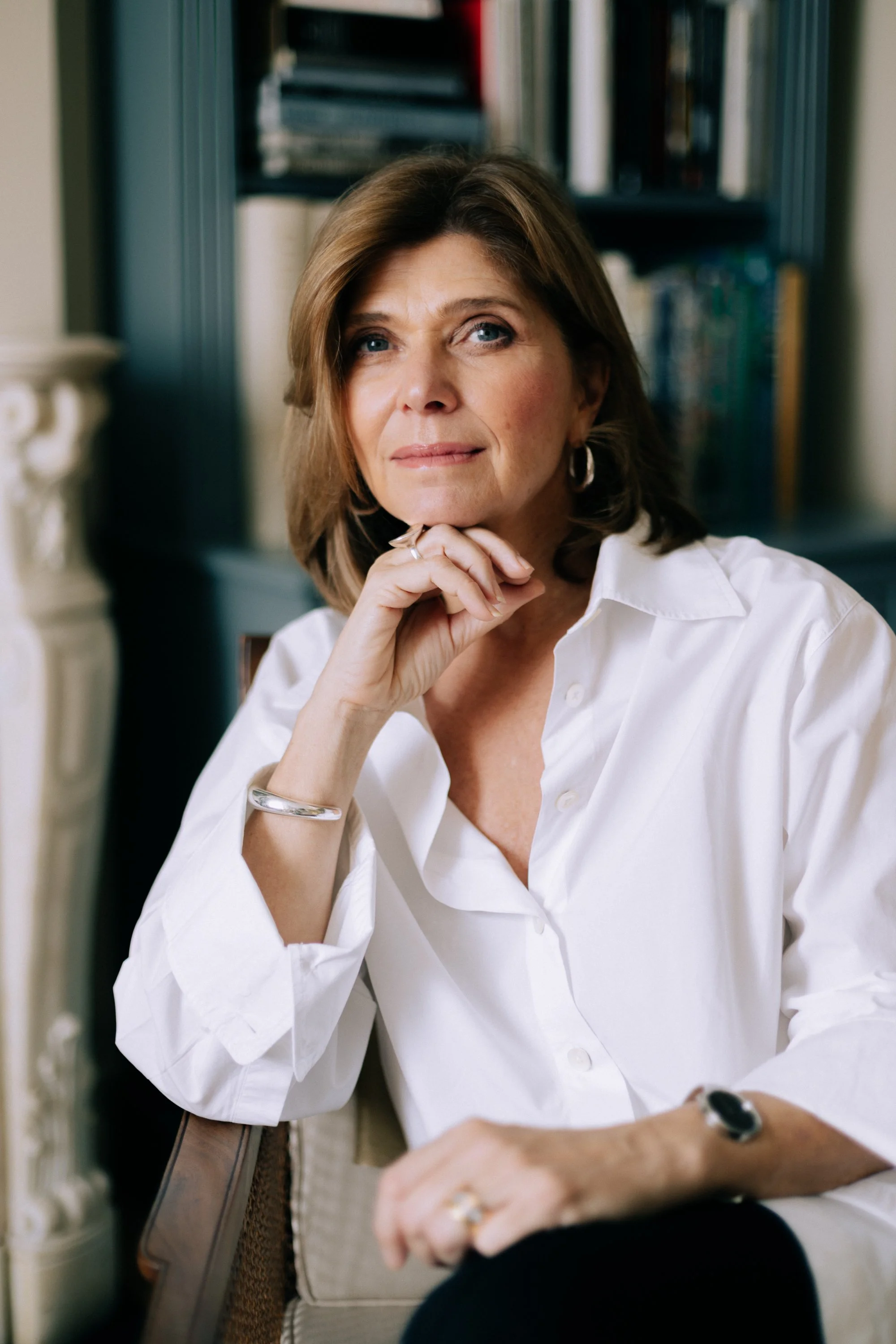 A middle-aged woman with shoulder-length brown hair and blue eyes, sitting in a chair with her chin resting on her hand, wearing a white button-up shirt, silver jewelry, and a thoughtful expression, with a bookshelf in the background.