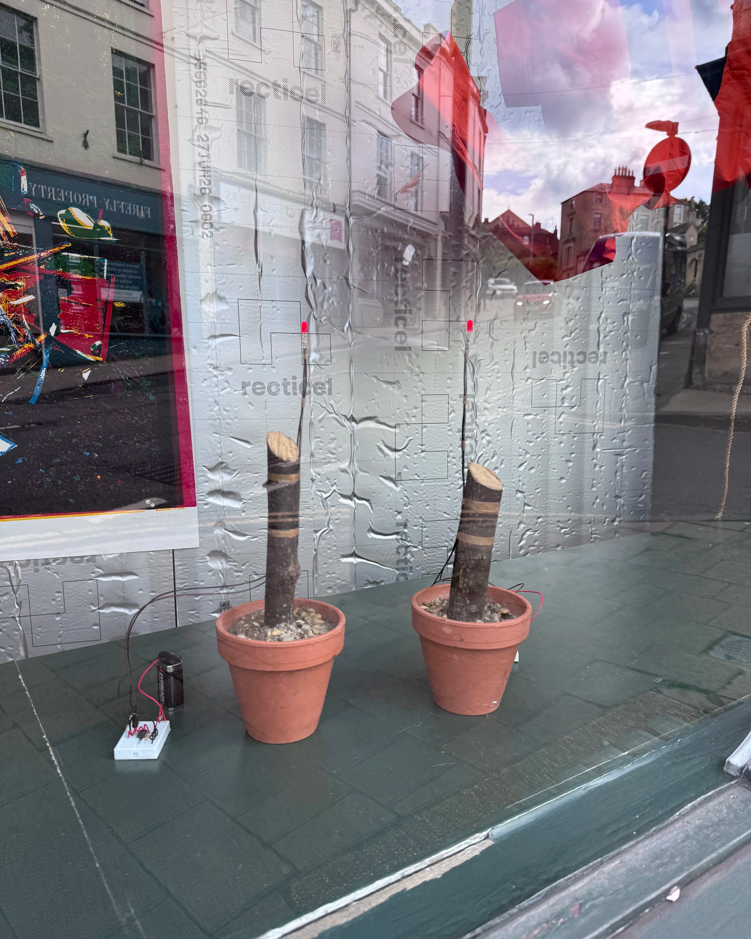 artworks: two terracotta plant pots filled with concrete and sawn off sticks with flashing red LED lights attached sit on a painted green window sill, in front of sheets of metallic insulation board with a brightly coloured linocut print attached